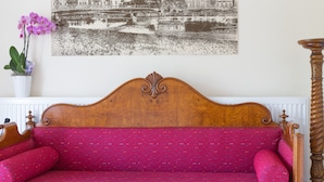 Un sofá de madera con asiento y cojines tapizados en rojo, situado frente a una pared con un cuadro de un paisaje urbano en blanco y negro.