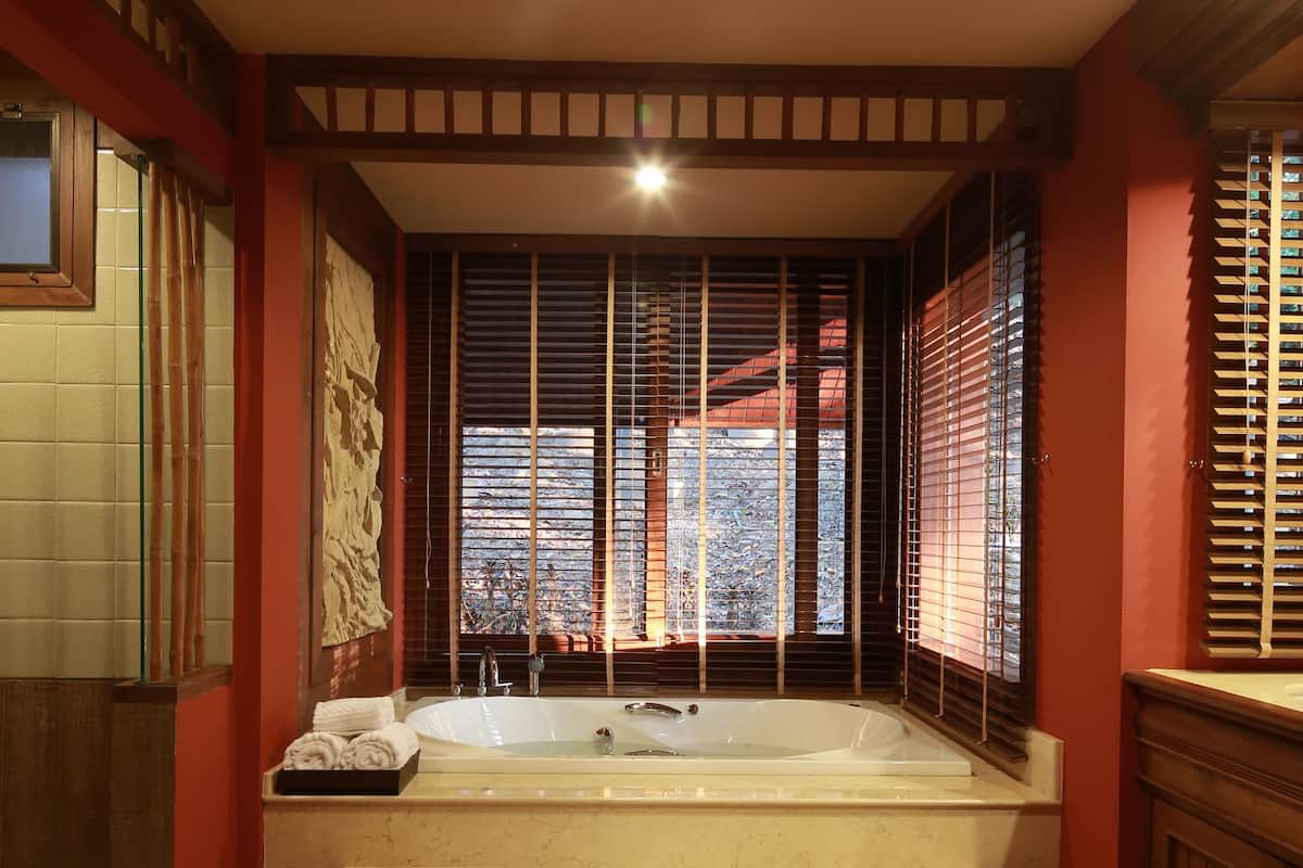 A room with a large bathtub, wooden beams on the ceiling, and a window with blinds.