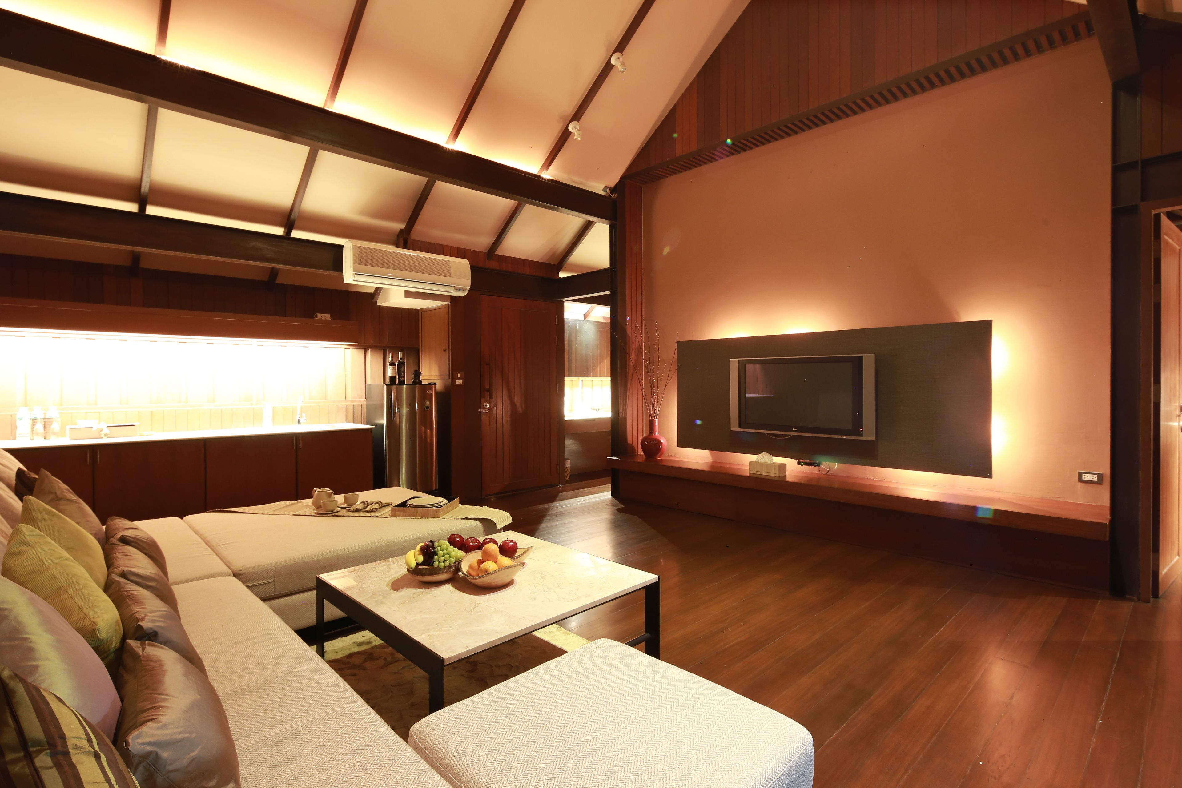 A spacious living area with a large sofa, a coffee table with fruit, a flat-screen TV mounted on the wall, and wooden flooring.