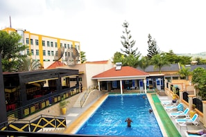 Ein Schwimmbecken mit Liegestühlen, einer überdachten Lounge und einem Gebäude mit gelb-weißen Wänden.