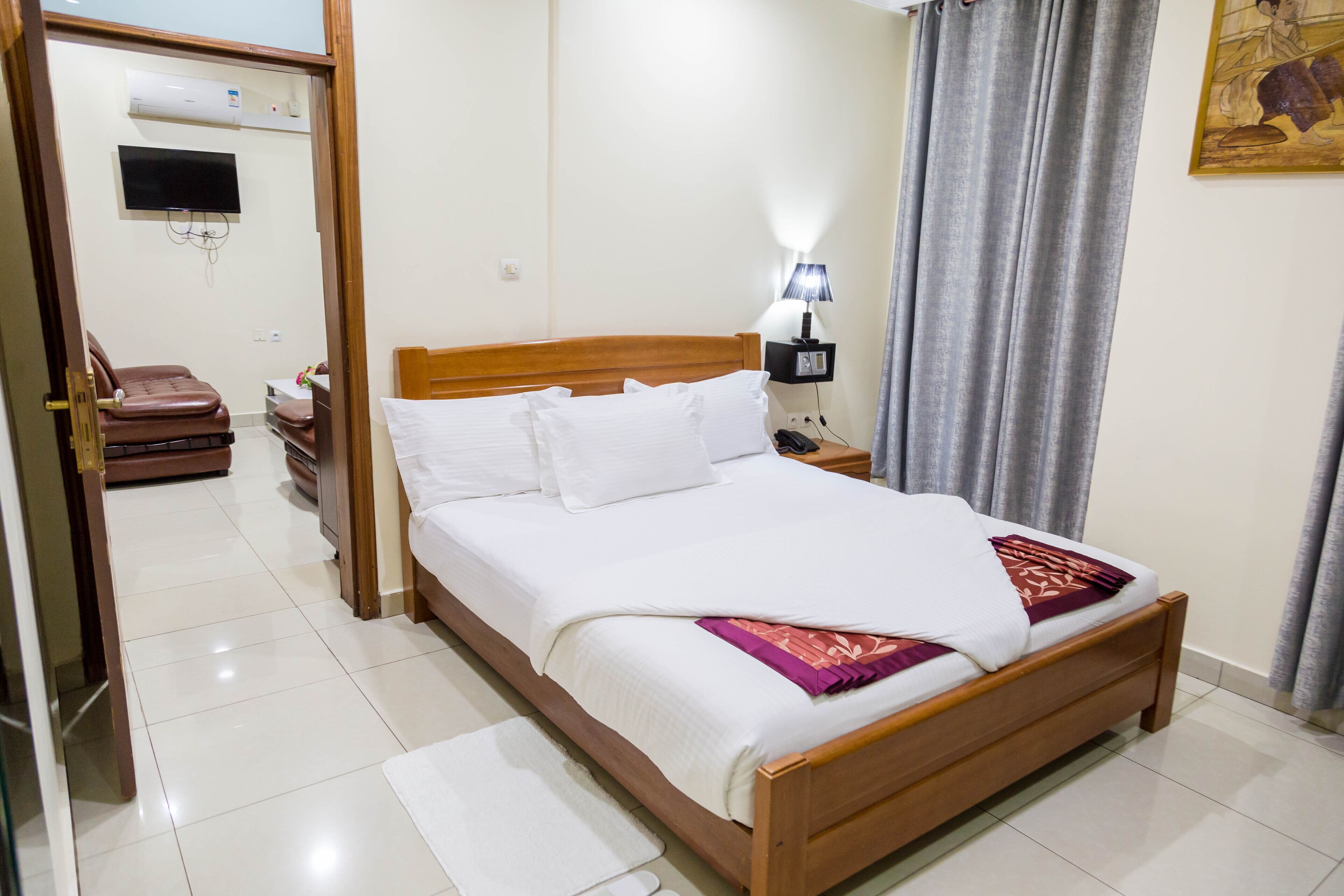 A neatly made bed with white linens and a purple throw, a bedside lamp, and a television mounted on the wall.