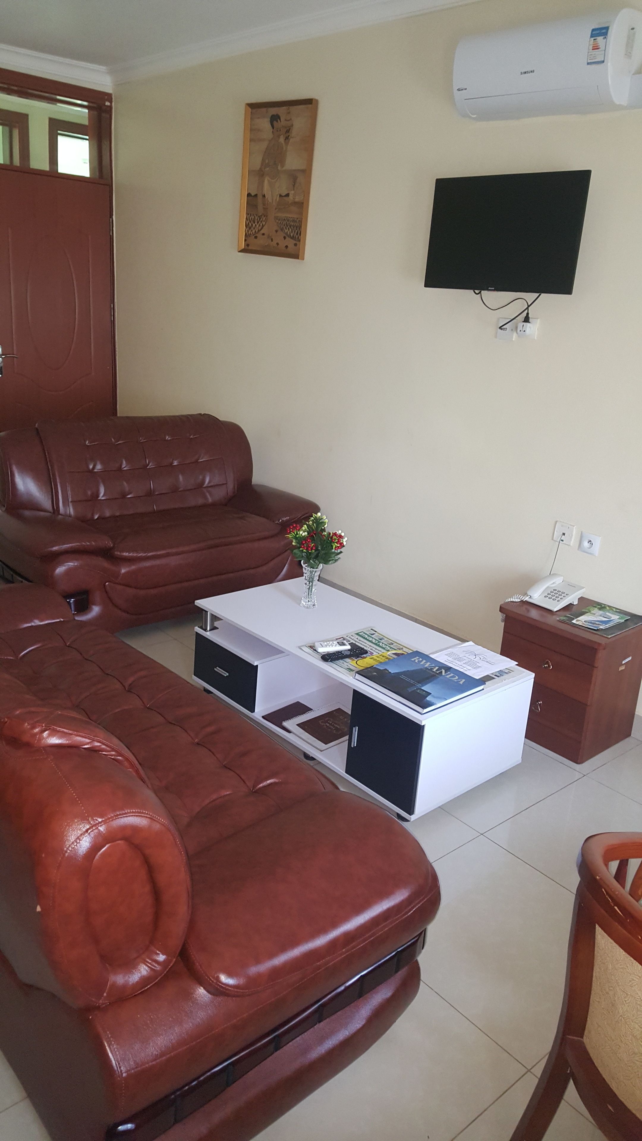 A living room with a brown leather sofa, a white coffee table with magazines and a vase of flowers, a television on the wall, and a wooden cabinet with a phone and papers.