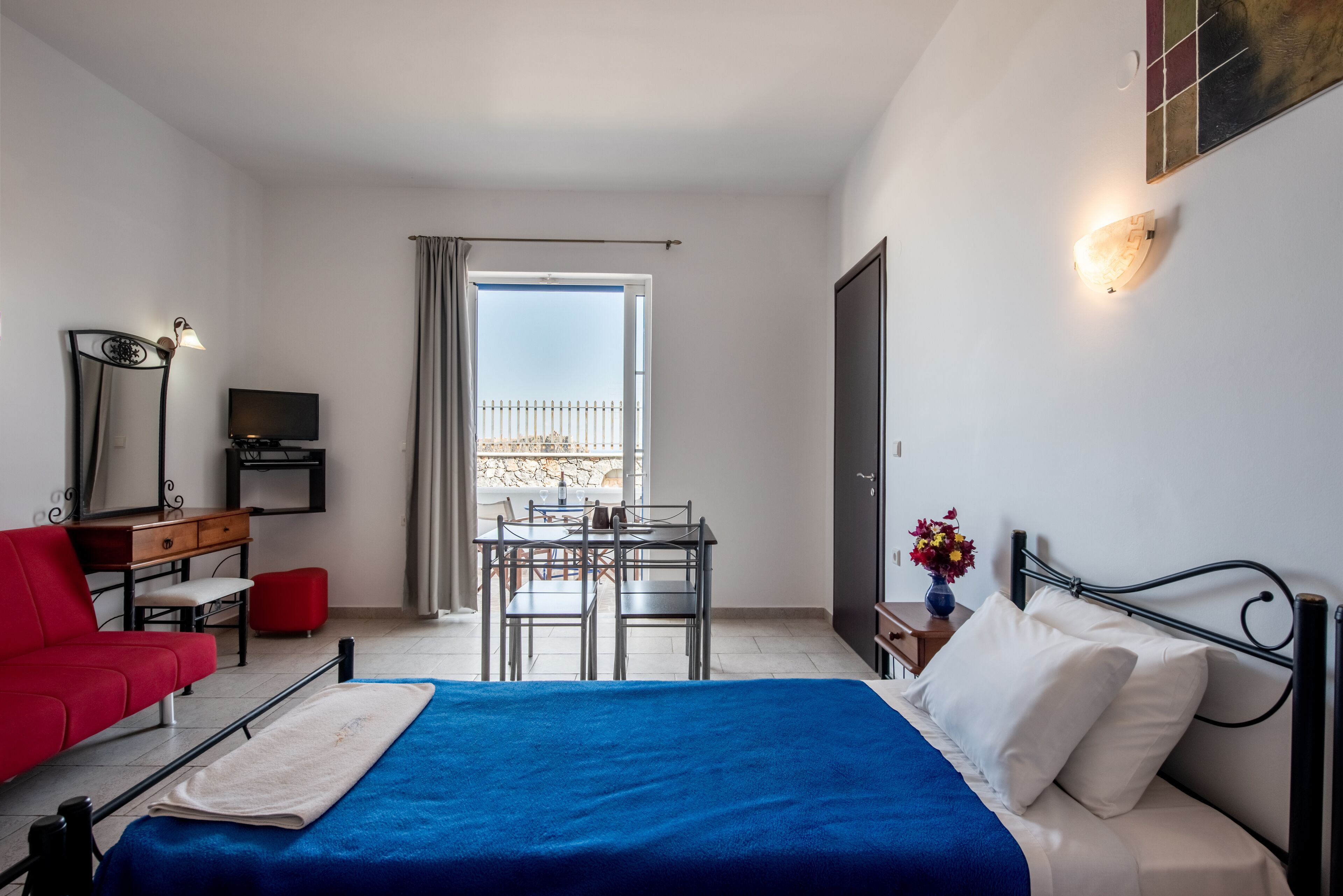 A hotel room with a bed, a red chair, a desk, a TV, and a balcony with a view.