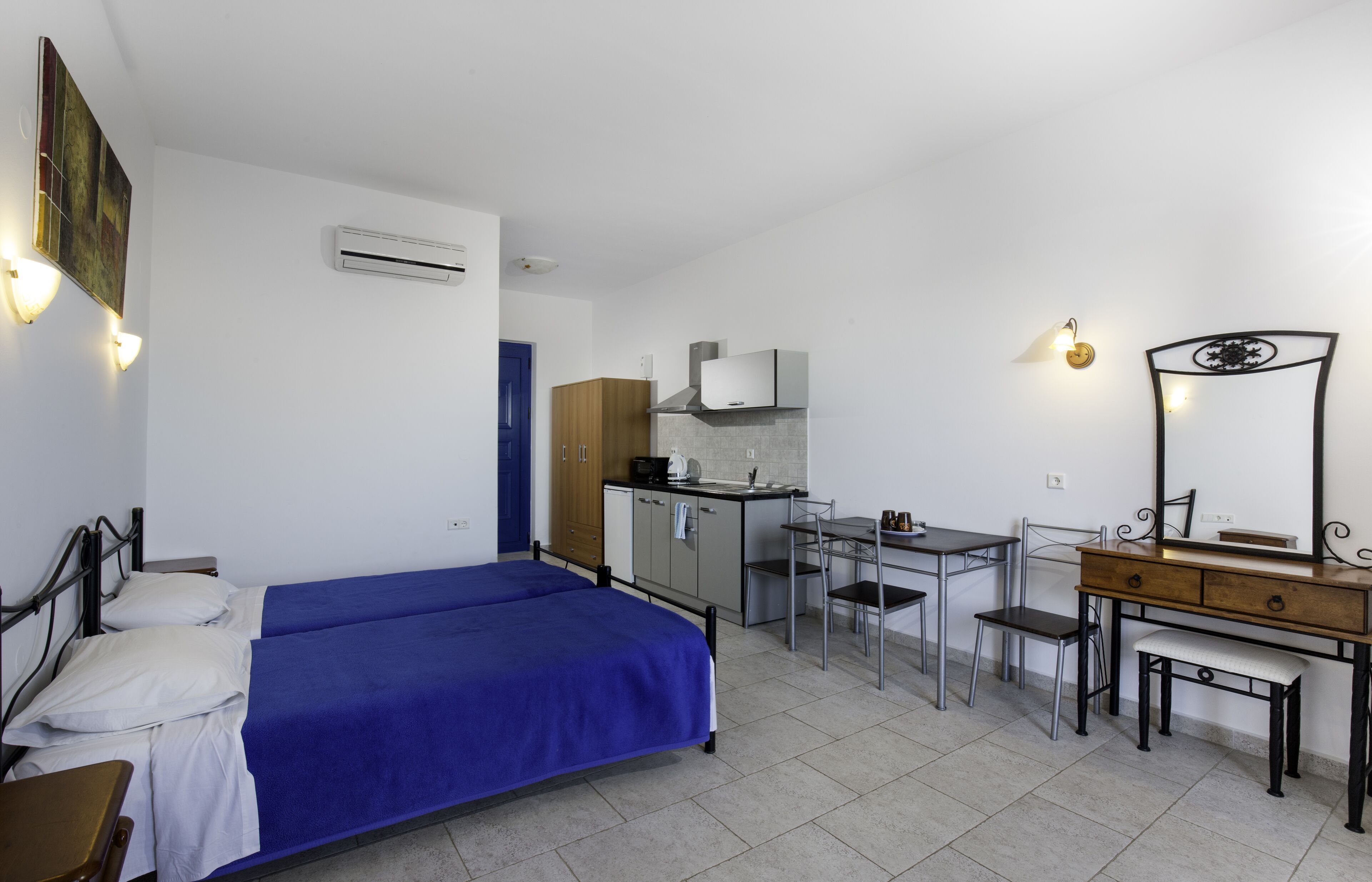 A compact room with a bed, a small kitchenette, a desk with a chair, and a vanity with a mirror.
