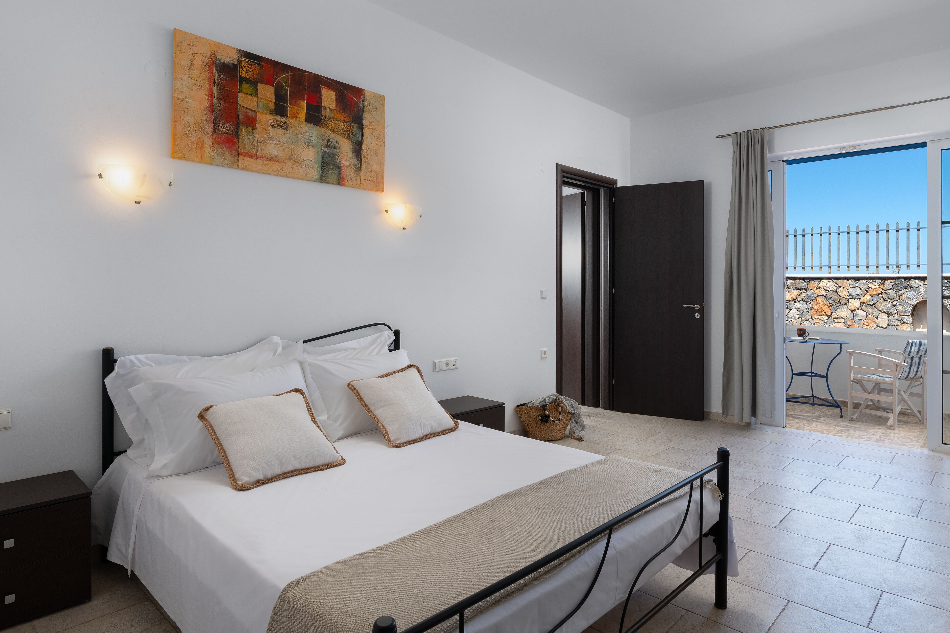 A bedroom with a bed, a painting, a nightstand, a chair, and a balcony with a stone wall.