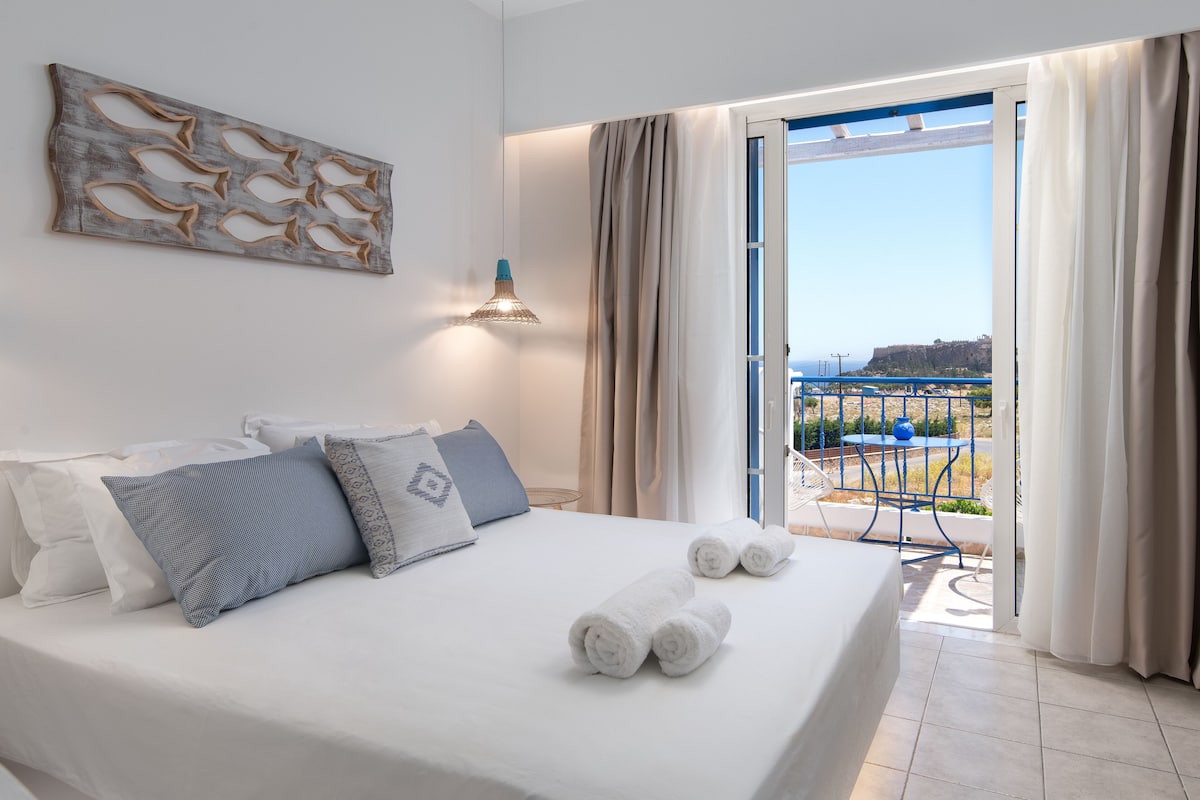 Habitación de hotel con cama, vista al mar y balcón con mesa y sillas.
