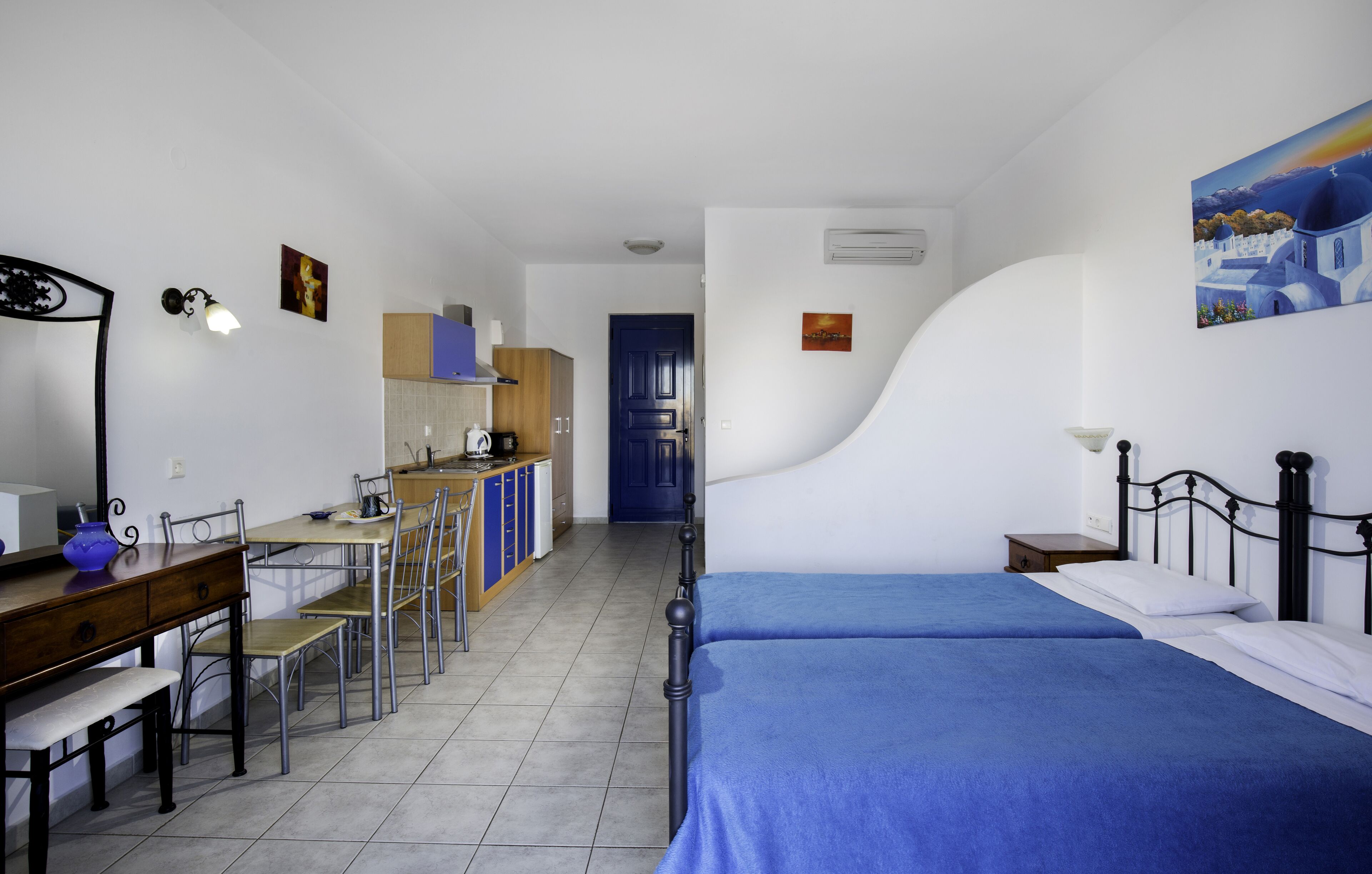A compact hotel room with a bed, a desk, and a chair. There is a kitchenette in the background.