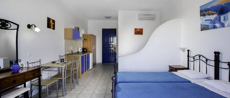 A compact hotel room with a bed, a desk, and a chair. There is a kitchenette in the background.