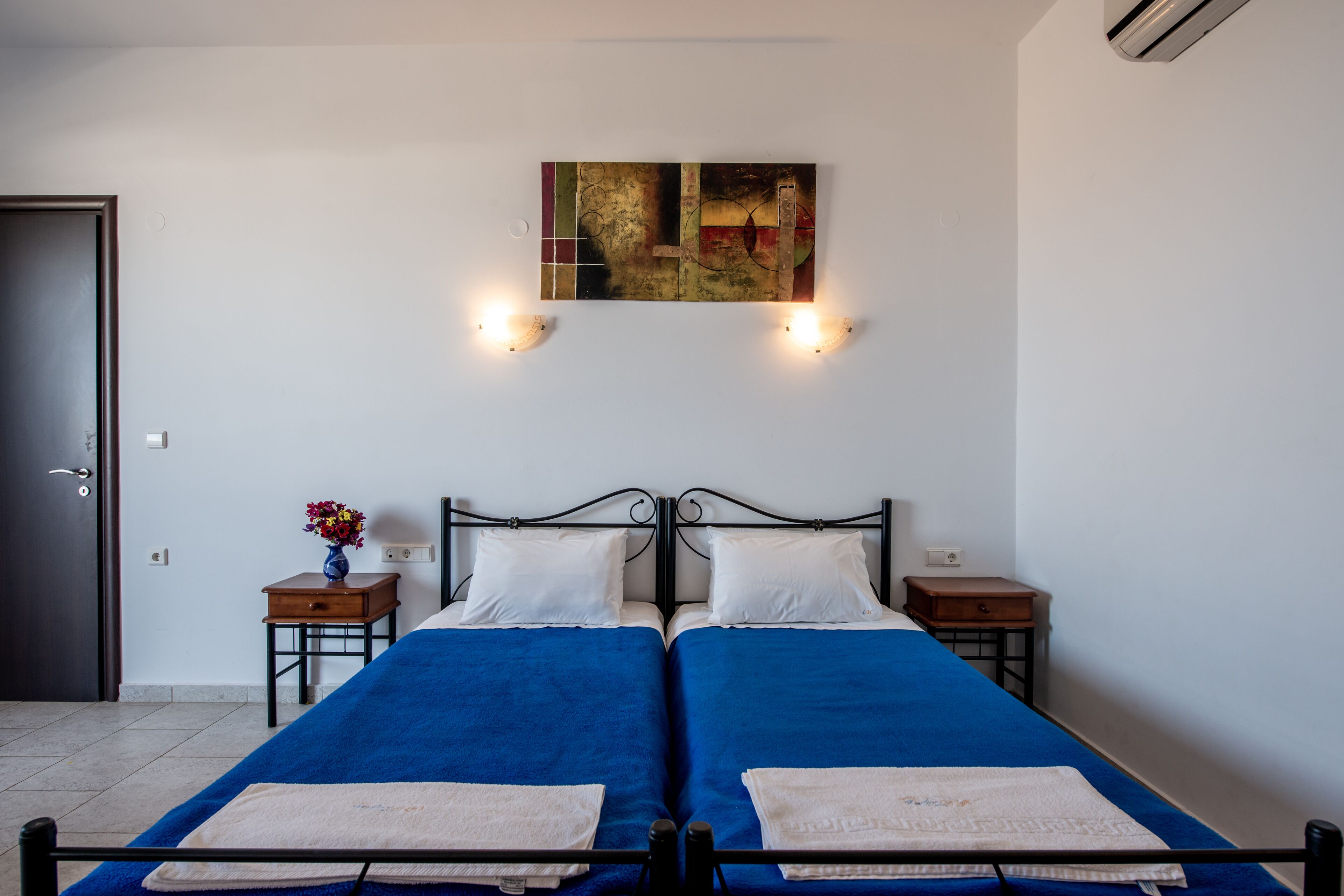 A double bed with blue bedding, two bedside tables, a wall-mounted artwork, and a door.