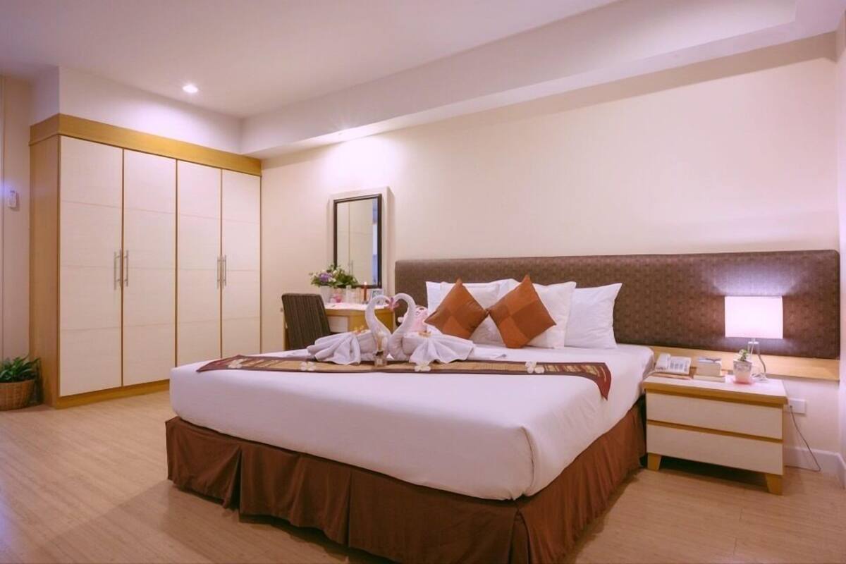 A hotel room with a large bed, a nightstand, a desk with a lamp, a wardrobe, and a mirror.