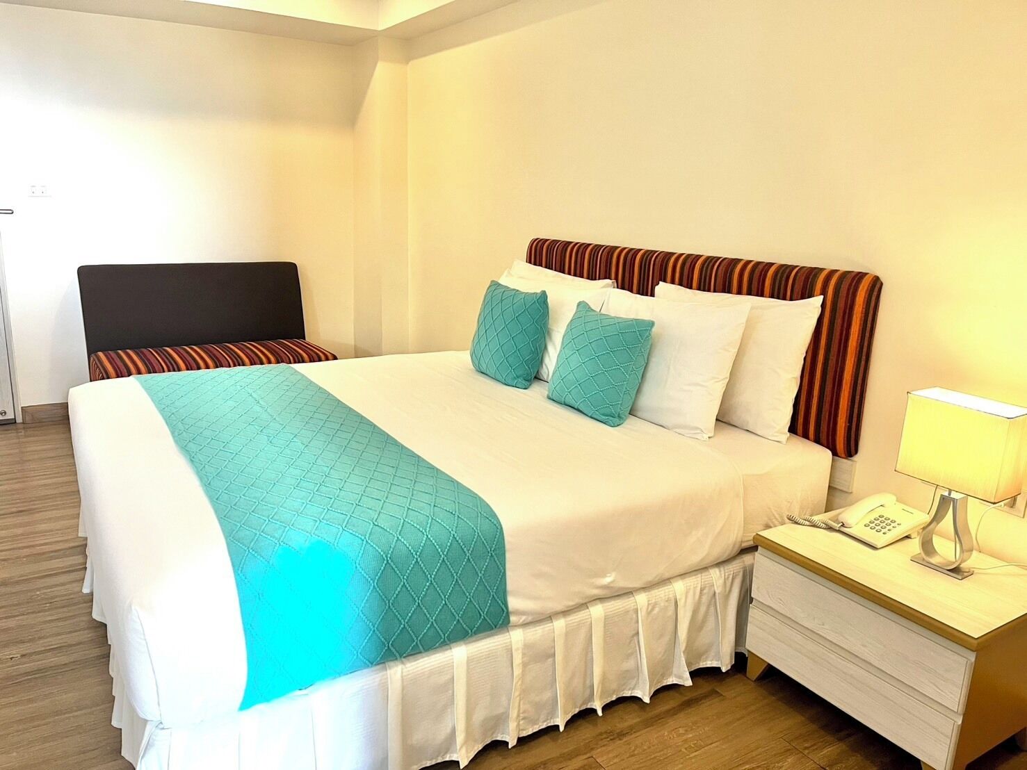 A neatly made bed with a white and teal bedspread, two teal pillows, and a striped headboard. There is a bedside table with a lamp and a phone.