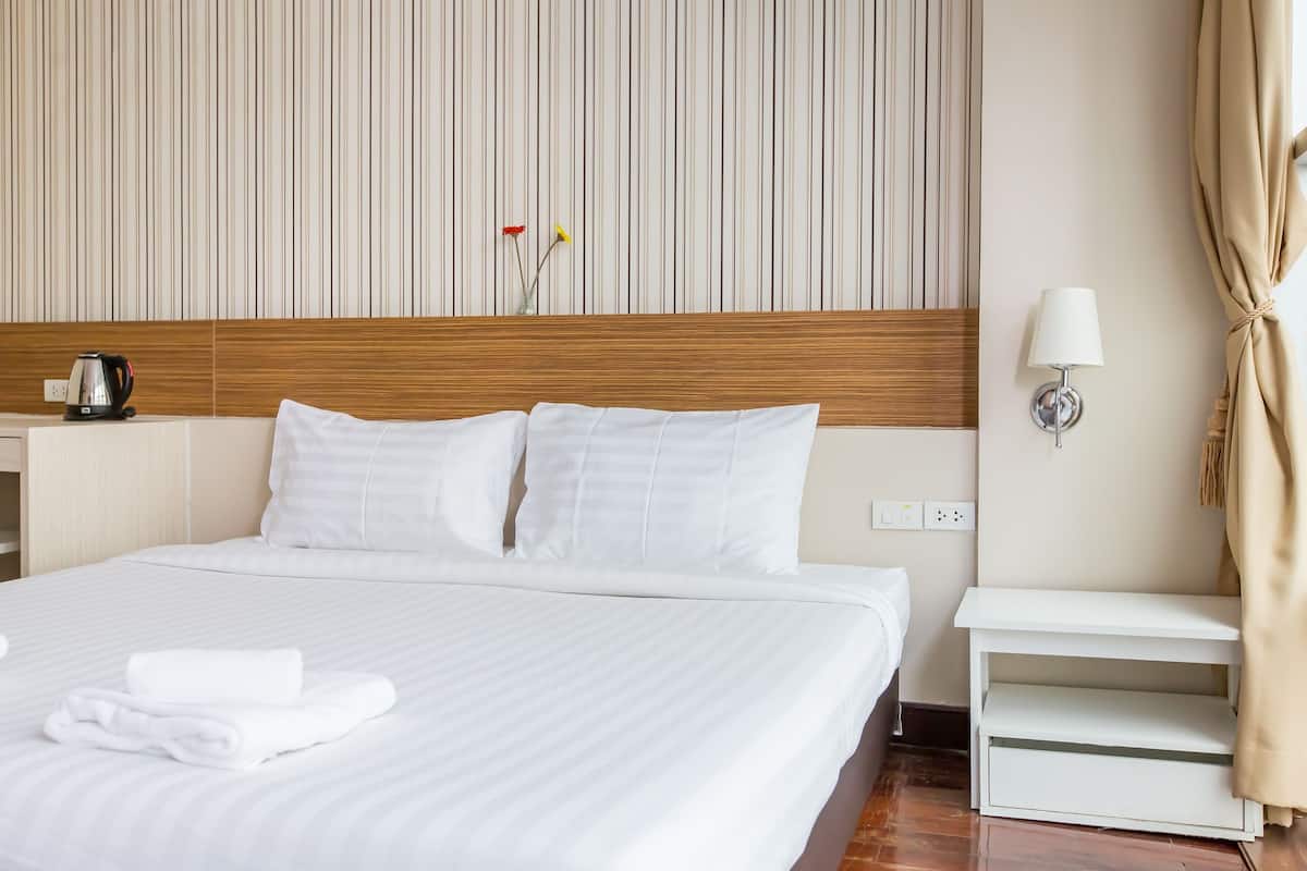 Habitación de hotel con una cama grande, ropa de cama blanca, cabecera de madera, mesita de noche con una lámpara y un florero con flores en la pared.