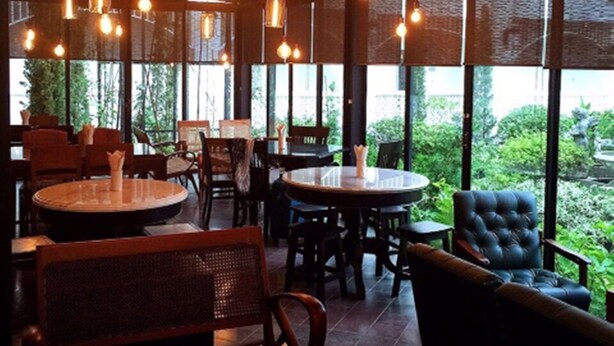 L’intérieur d’un restaurant présente des tables et des chaises rondes, des luminaires suspendus et de grandes fenêtres qui offrent une vue sur la verdure environnante.