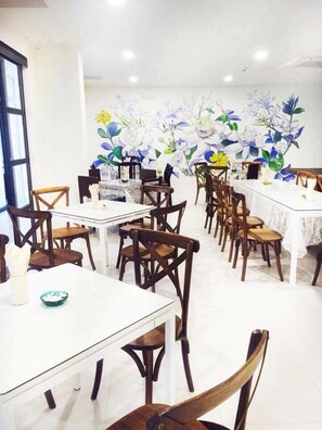 A dining area with multiple tables and chairs, a floral mural on the wall, and a window on the left. - Koranaree Courtyard Boutique Hotel (Nakhon Ratchasima)