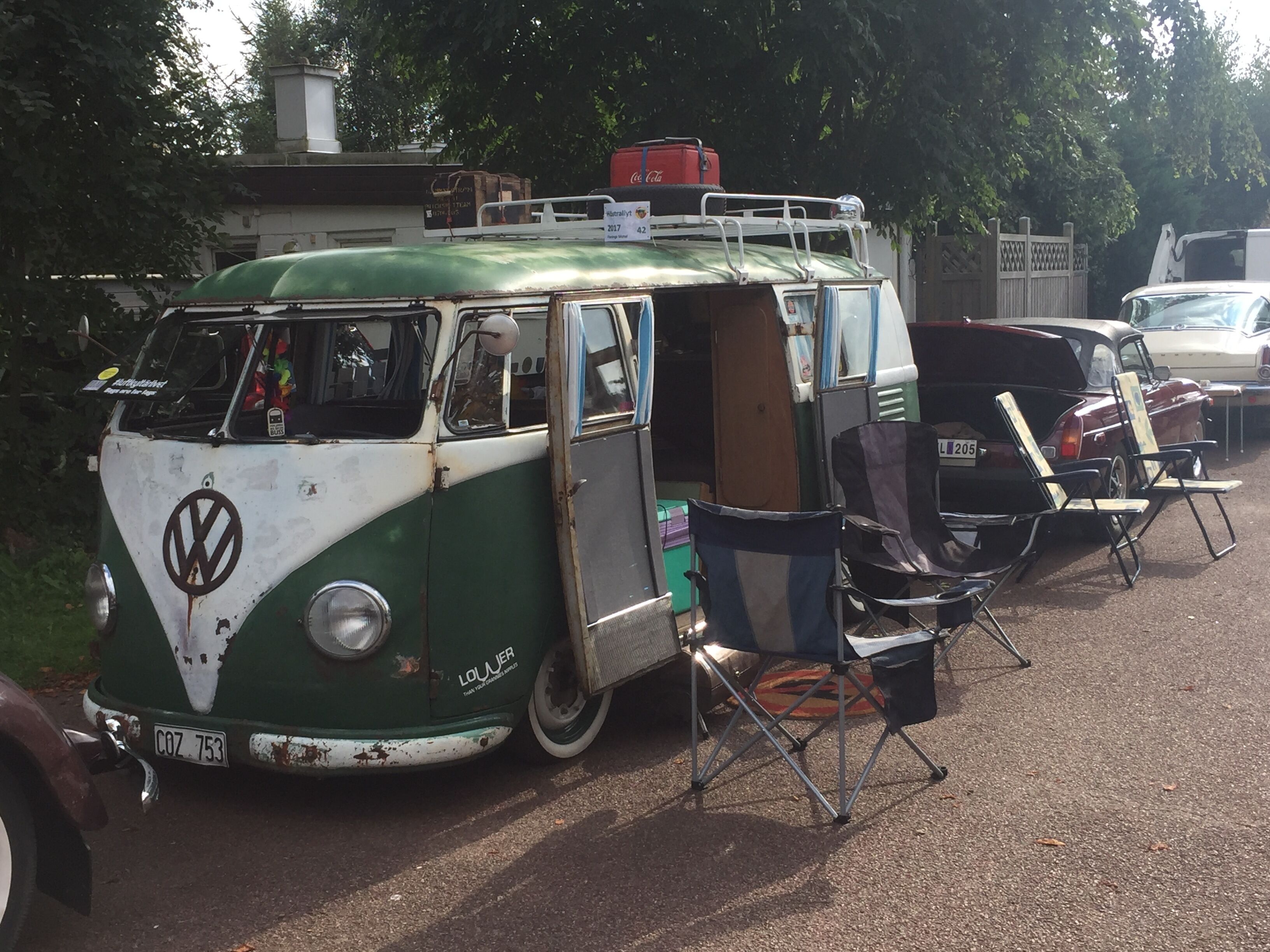 A vintage VW camper van parked on a street with a green and white exterior, a ladder on the roof, and a license plate reading COZ 753.