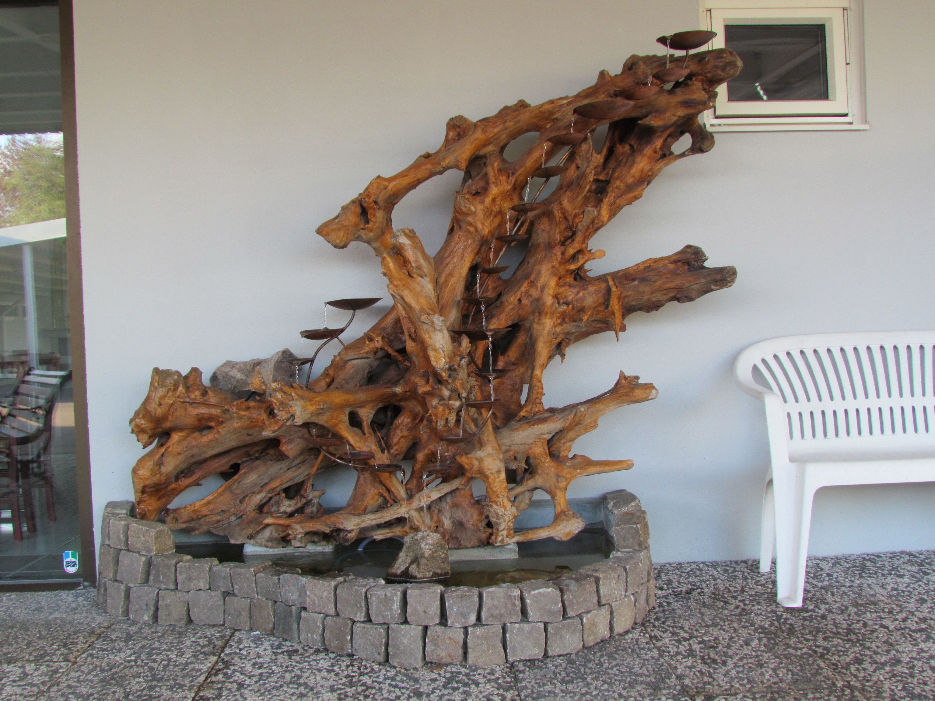 Una gran escultura de madera en una jardinera, una silla de plástico blanco y una ventana con vistas al exterior.
