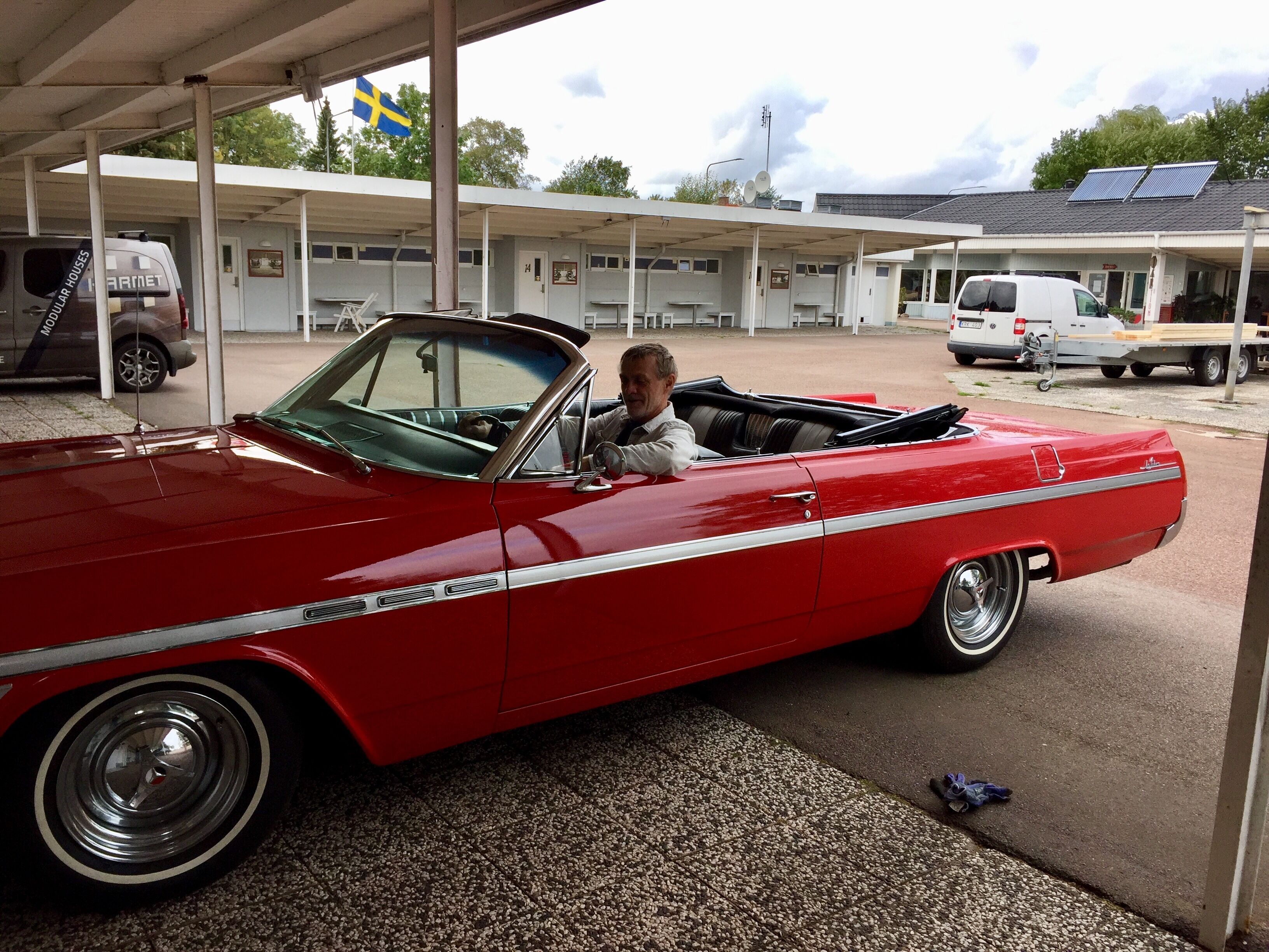 En röd cabriolet stod parkerad under ett tak med en svensk flagga, bredvid en byggnad med flera rum, och på taket satt en solpanel.
