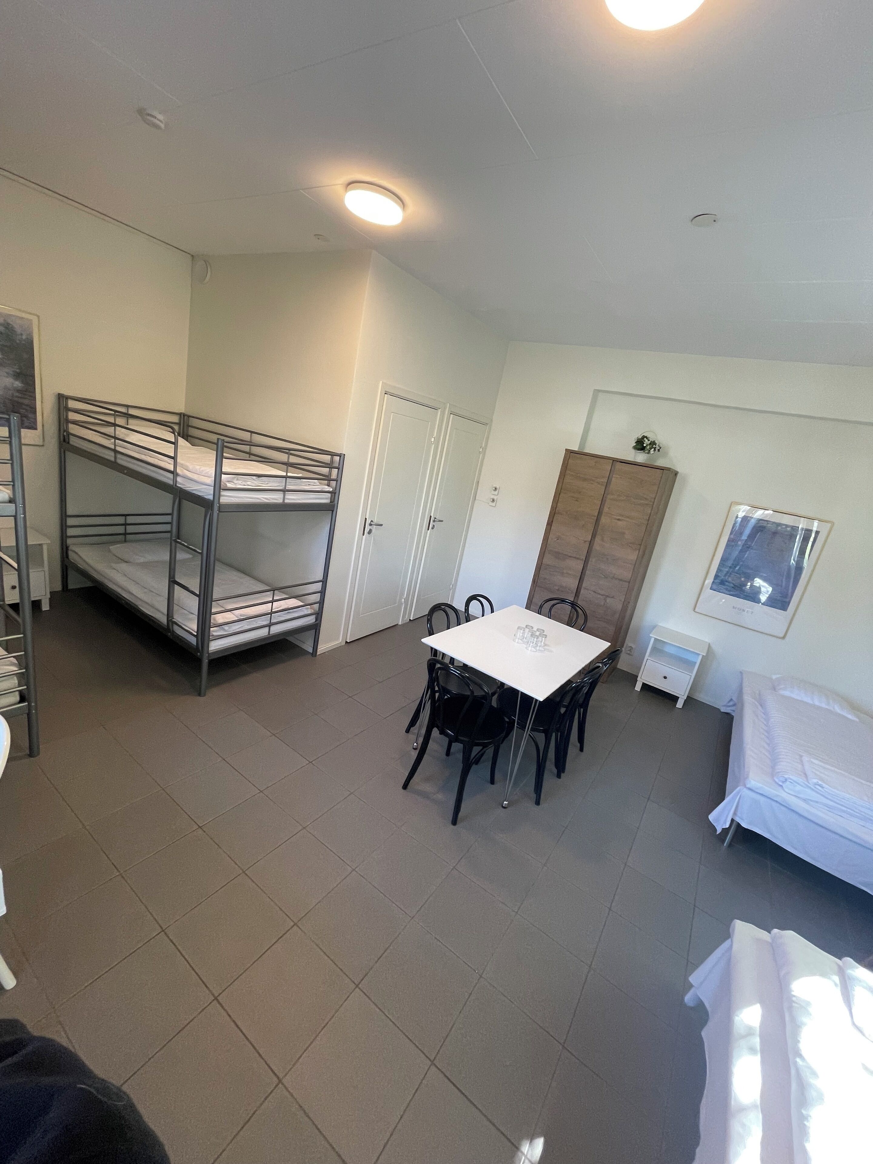 A dormitory room with bunk beds, a dining table with chairs, a wardrobe, and a small cabinet.