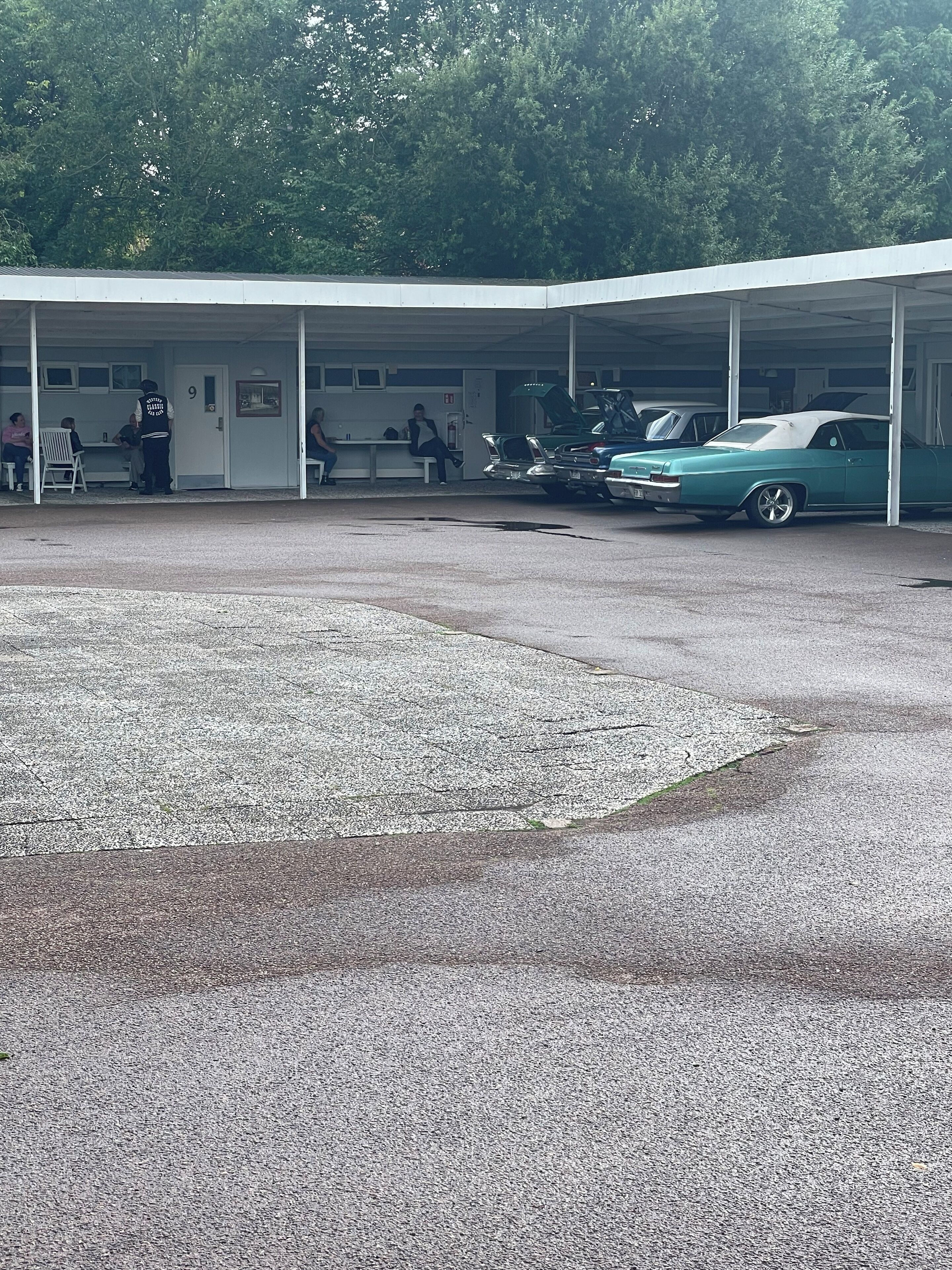 Un área de estacionamiento techada con un coche clásico, un edificio de techo plano y personas sentadas en bancos.