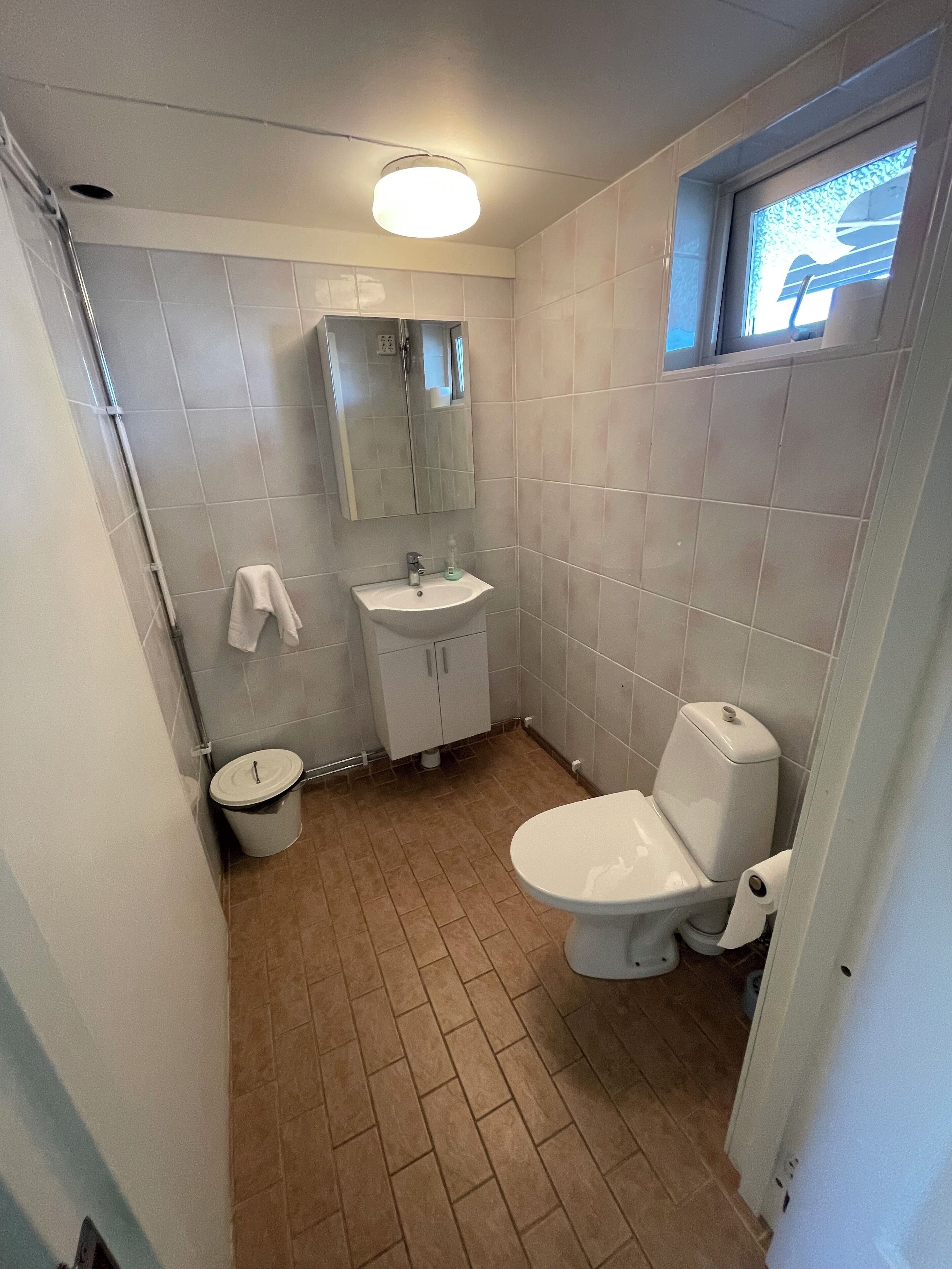 Un baño pequeño y funcional con un lavamanos, espejo y retrete blancos. Hay una ventana sobre el retrete y una toalla colgada en la pared.