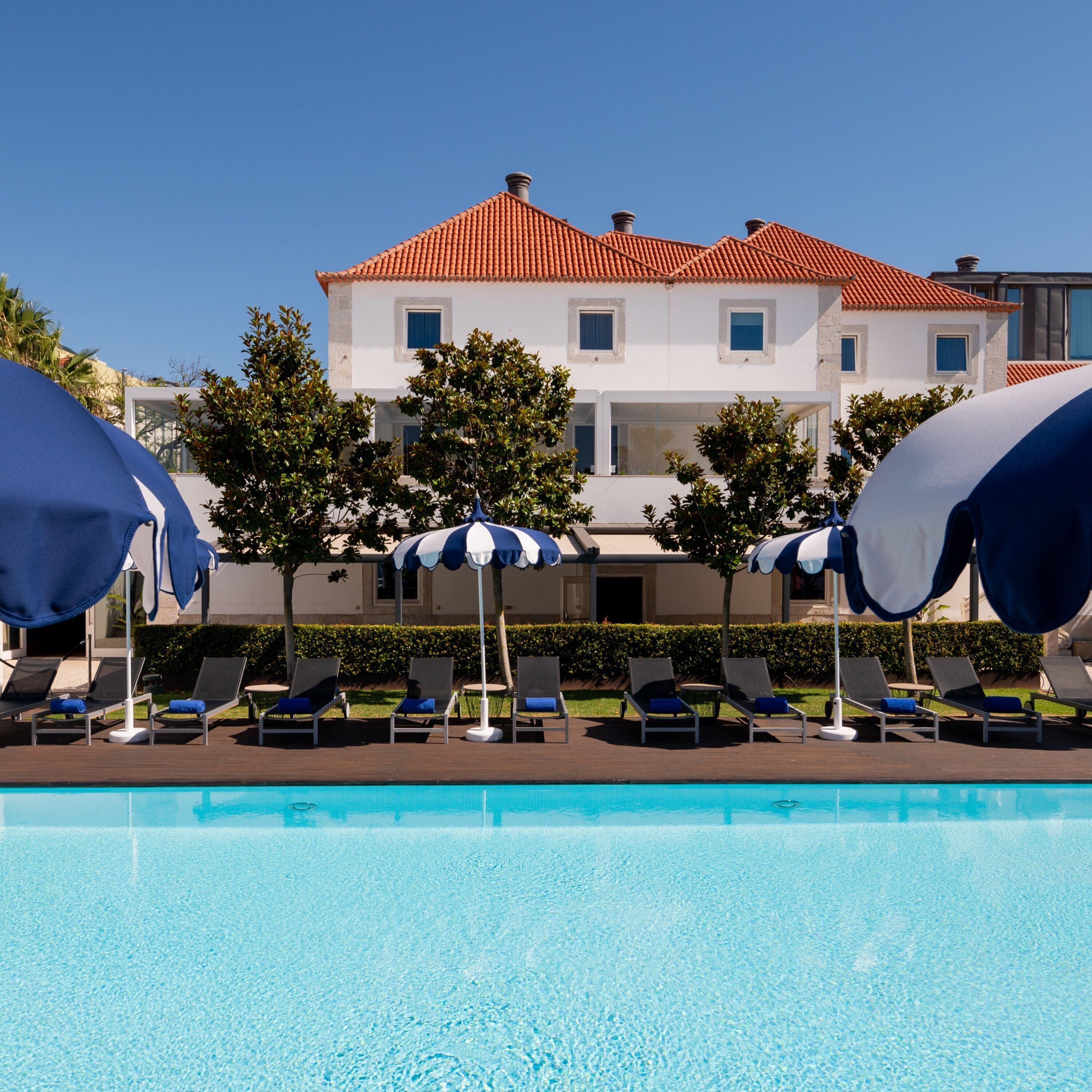 A pool area with lounge chairs and umbrellas, a clear blue sky, and a building with a red-tiled roof in the background.