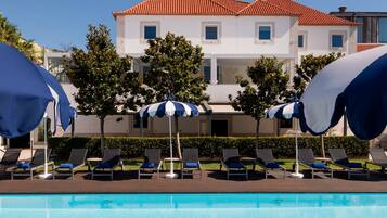 A pool area with lounge chairs and umbrellas, a clear blue sky, and a building with a red-tiled roof in the background.