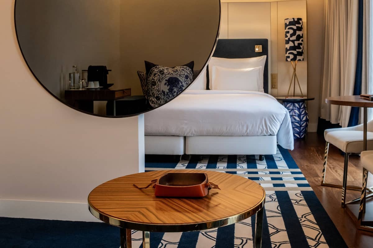A hotel room with a large mirror, a bed with white linens, a small table with a red bag, and a blue and white patterned rug.