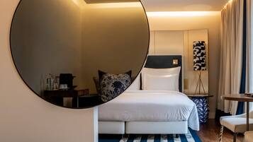 Quarto de hotel com um espelho grande, uma cama com lençóis brancos, uma mesa pequena com uma bolsa vermelha e um tapete com padrão azul e branco.
