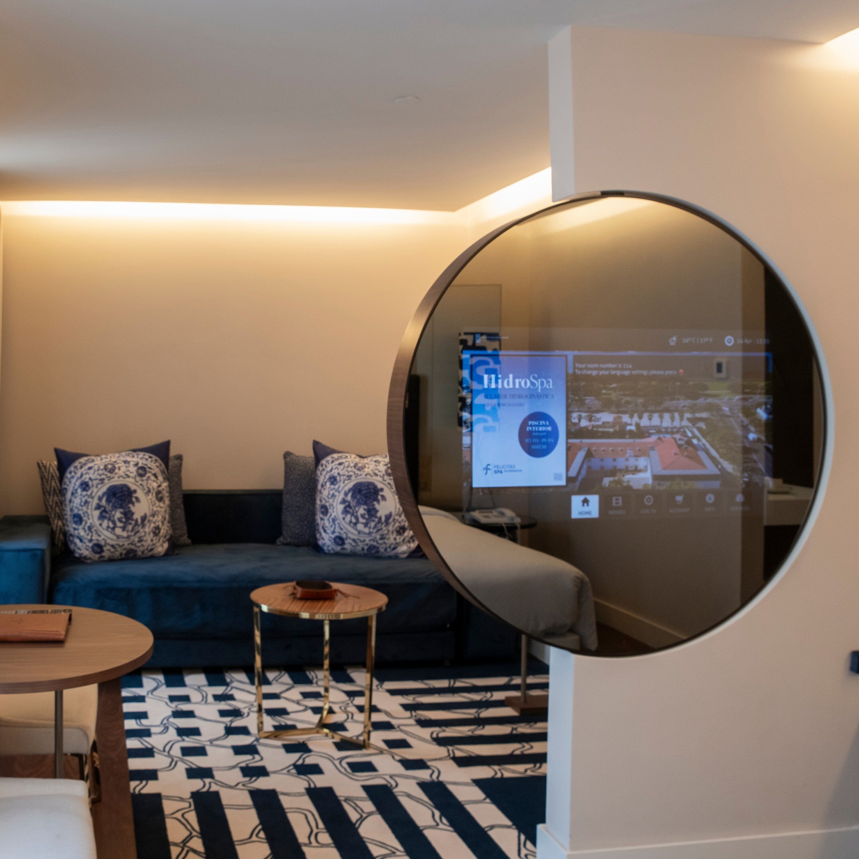 Una sala moderna con un espejo redondo que refleja un espacio con un sofá azul, cojines estampados, una mesita de madera y una alfombra con diseño.