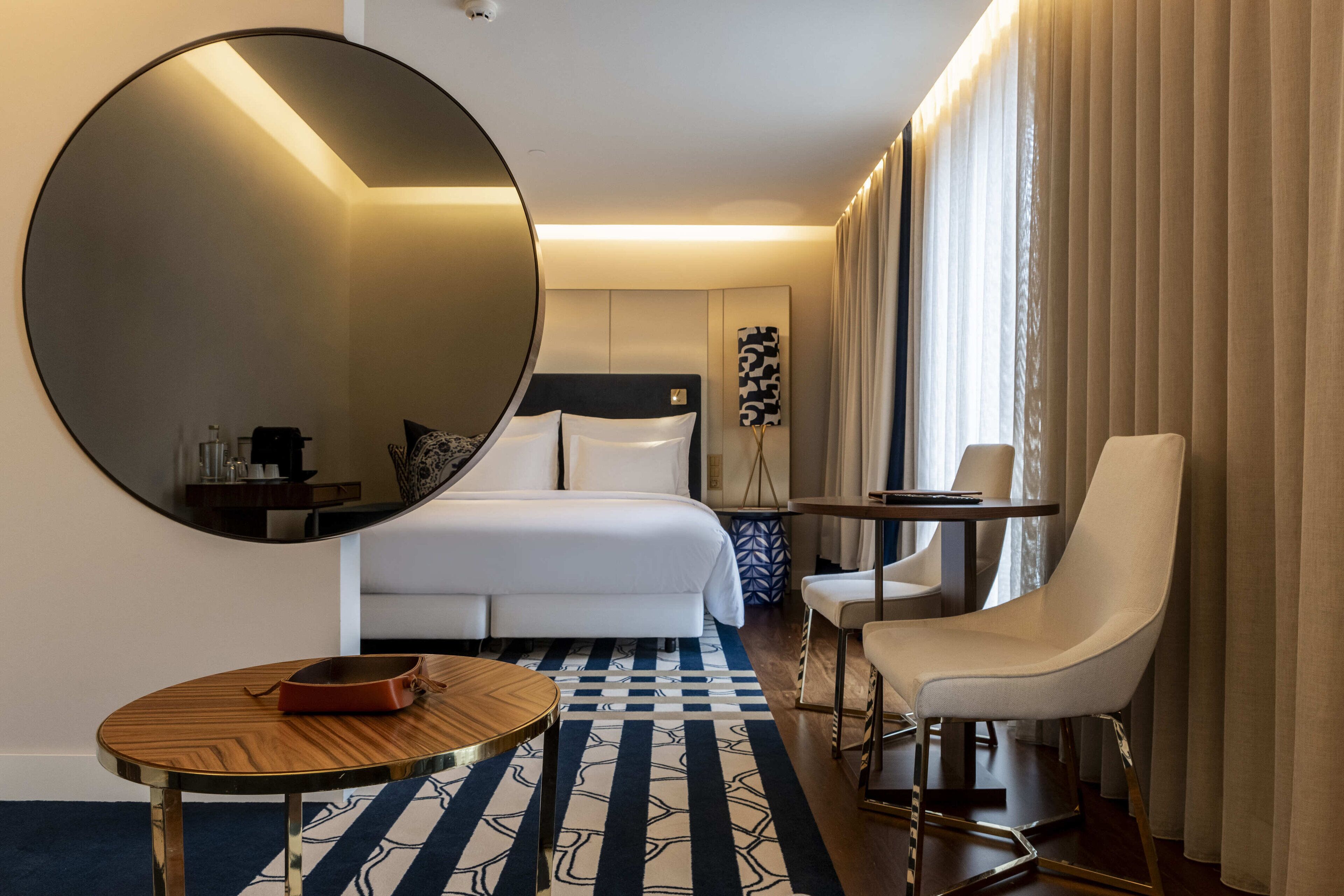 Ein Hotelzimmer mit einem großen runden Spiegel, einem Bett mit weißer Bettwäsche, einem kleinen runden Tisch mit einer braunen Ablage, einem weißen Sessel und einem gemusterten Teppich.