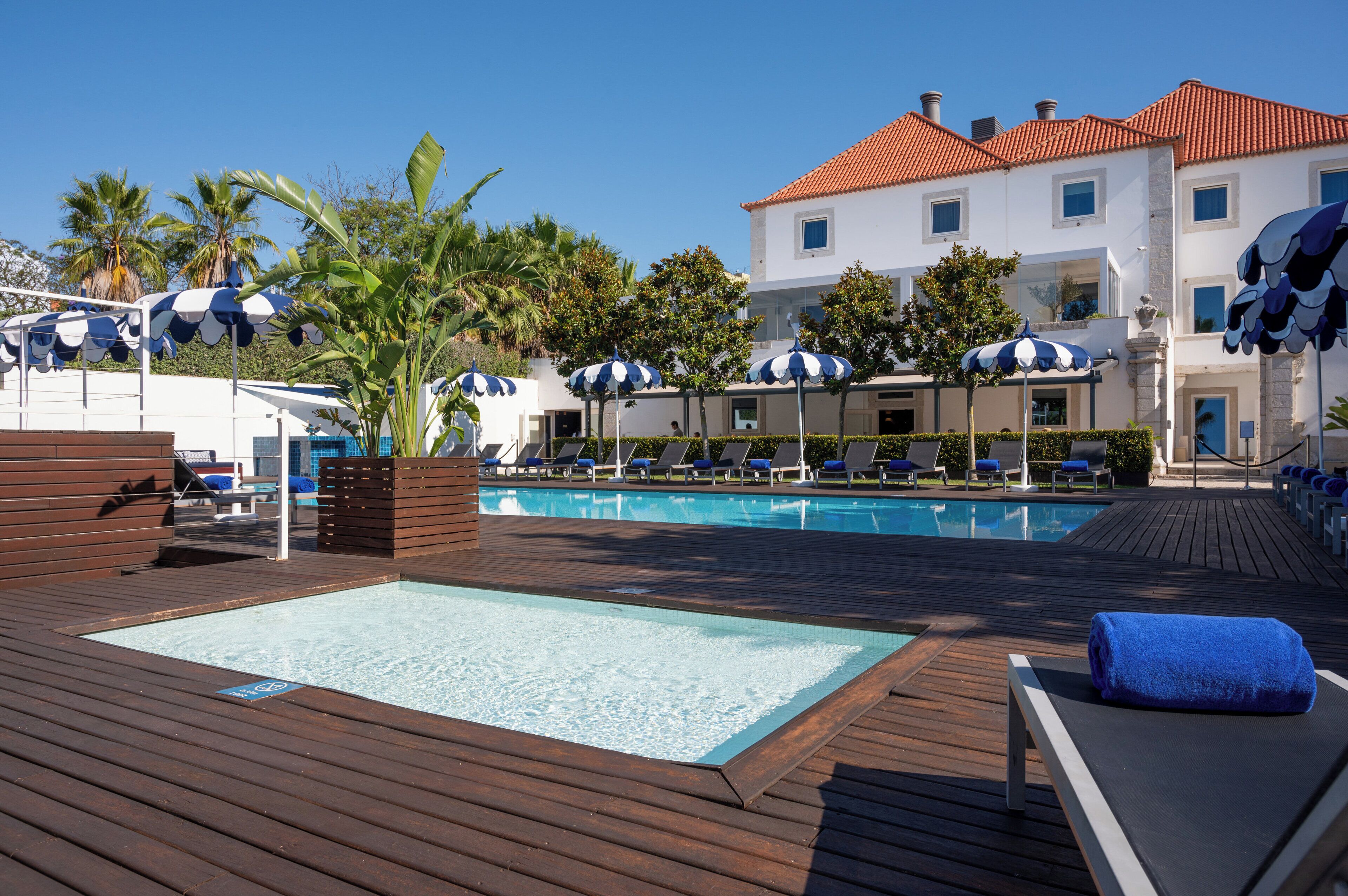 Un área de piscina con sillones de descanso y sombrillas, una terraza de madera y un edificio con tejado de tejas rojas.