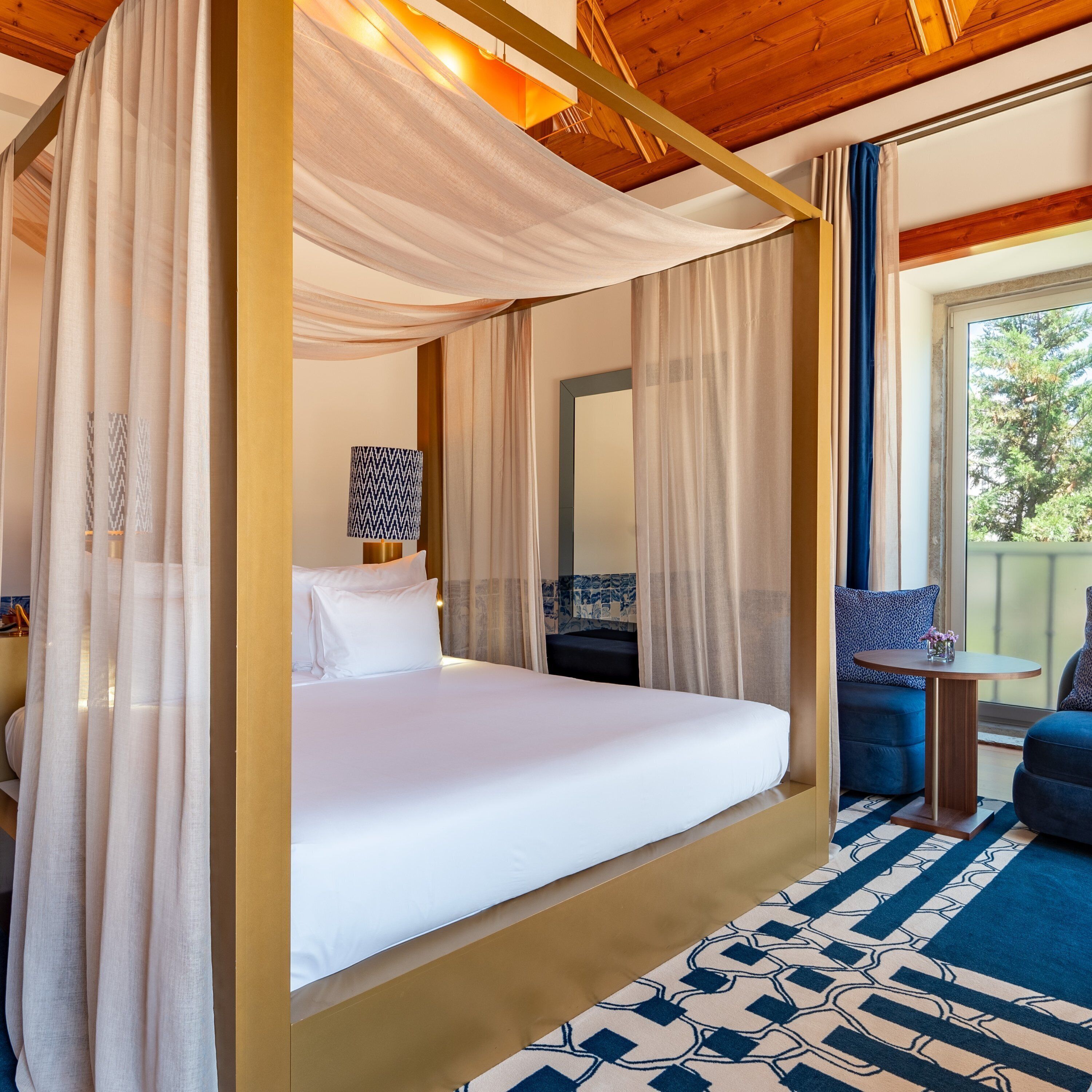 A four-poster bed with white linens, a bedside table with a lamp, and a view of the outdoors through a window.