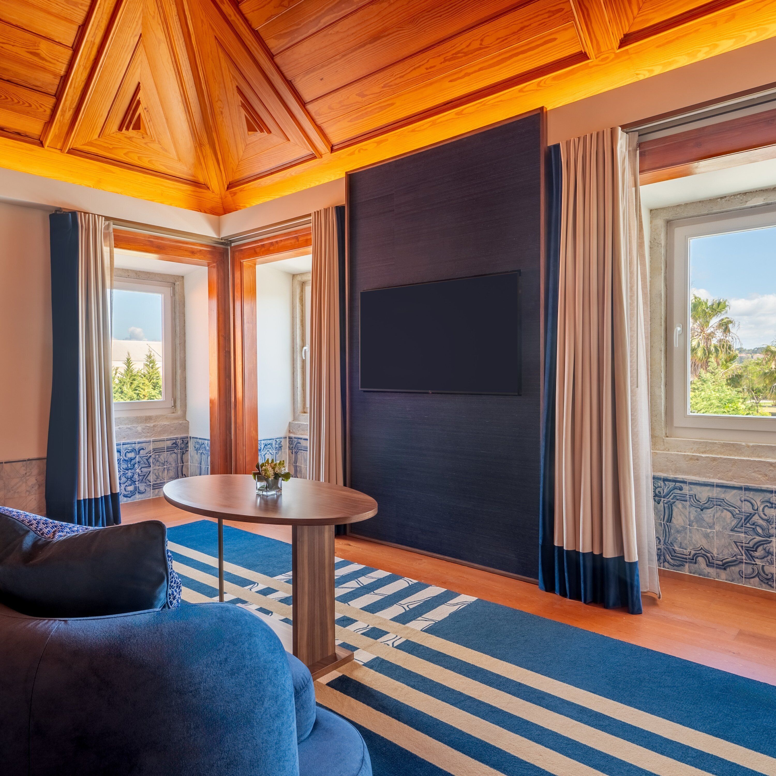 A room with a wooden ceiling, a round table with a vase, a sofa, and a large window with curtains.