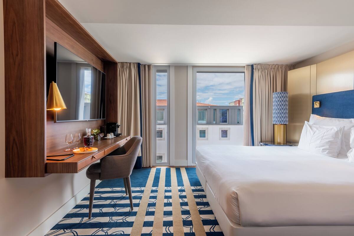 A hotel room with a bed, a desk with a chair, a large window, and a view of buildings outside.