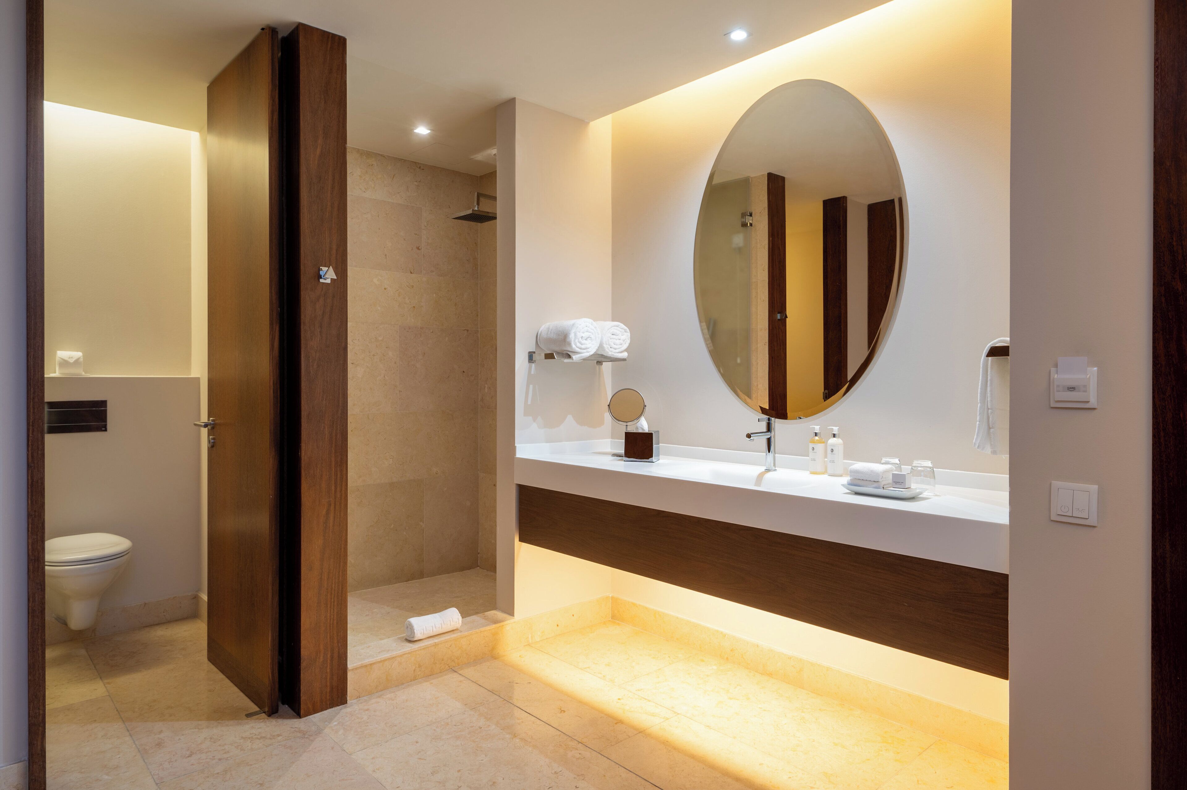 A modern bathroom with a walk-in shower, a large oval mirror, and a vanity with toiletries.