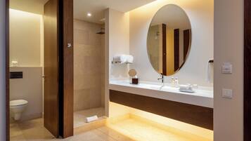 A modern bathroom with a walk-in shower, a large oval mirror, and a vanity with toiletries.