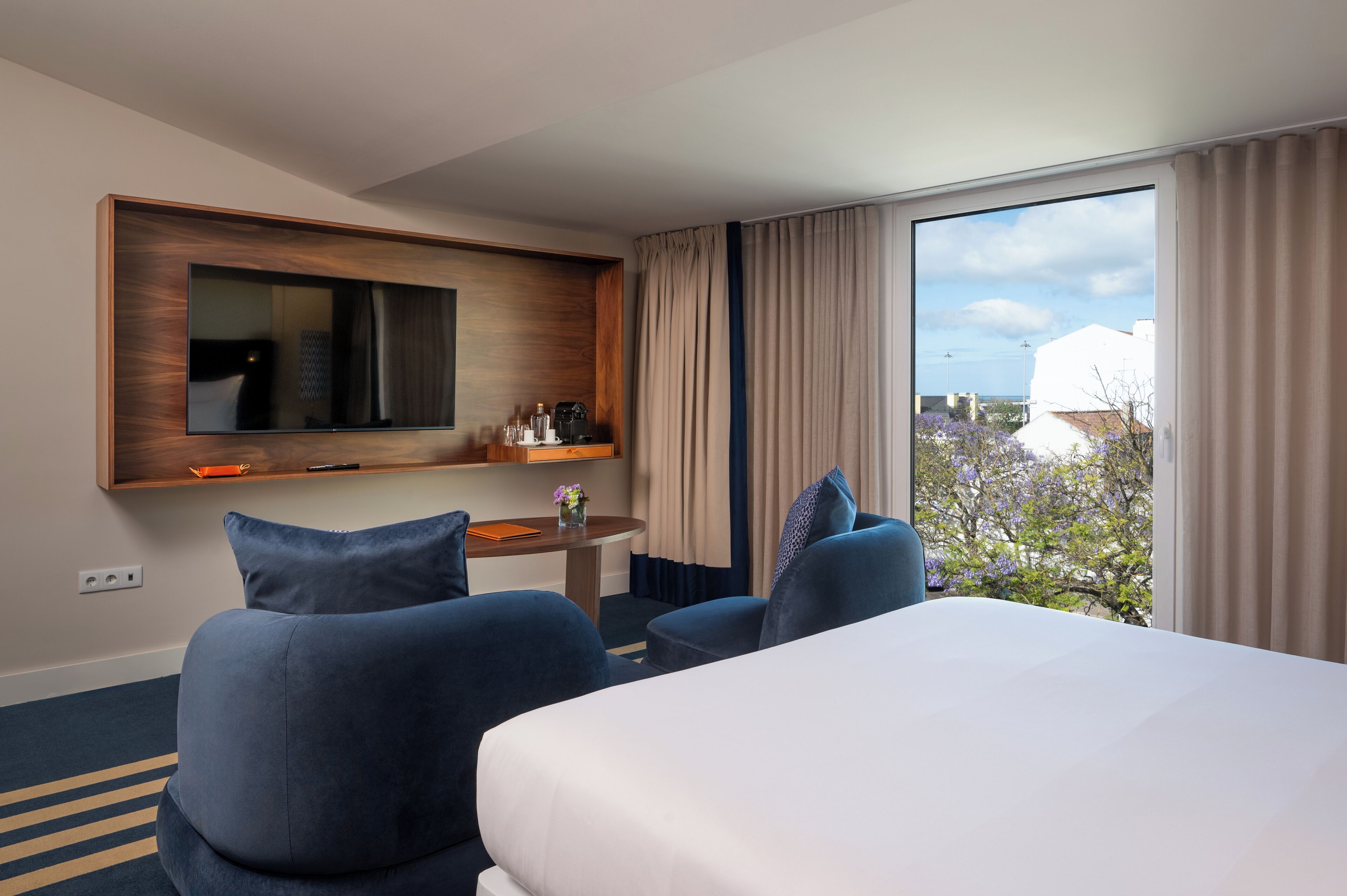 A hotel room with a bed, two armchairs, a small table with a vase of flowers, a TV mounted on a wooden panel, and a large window with curtains.