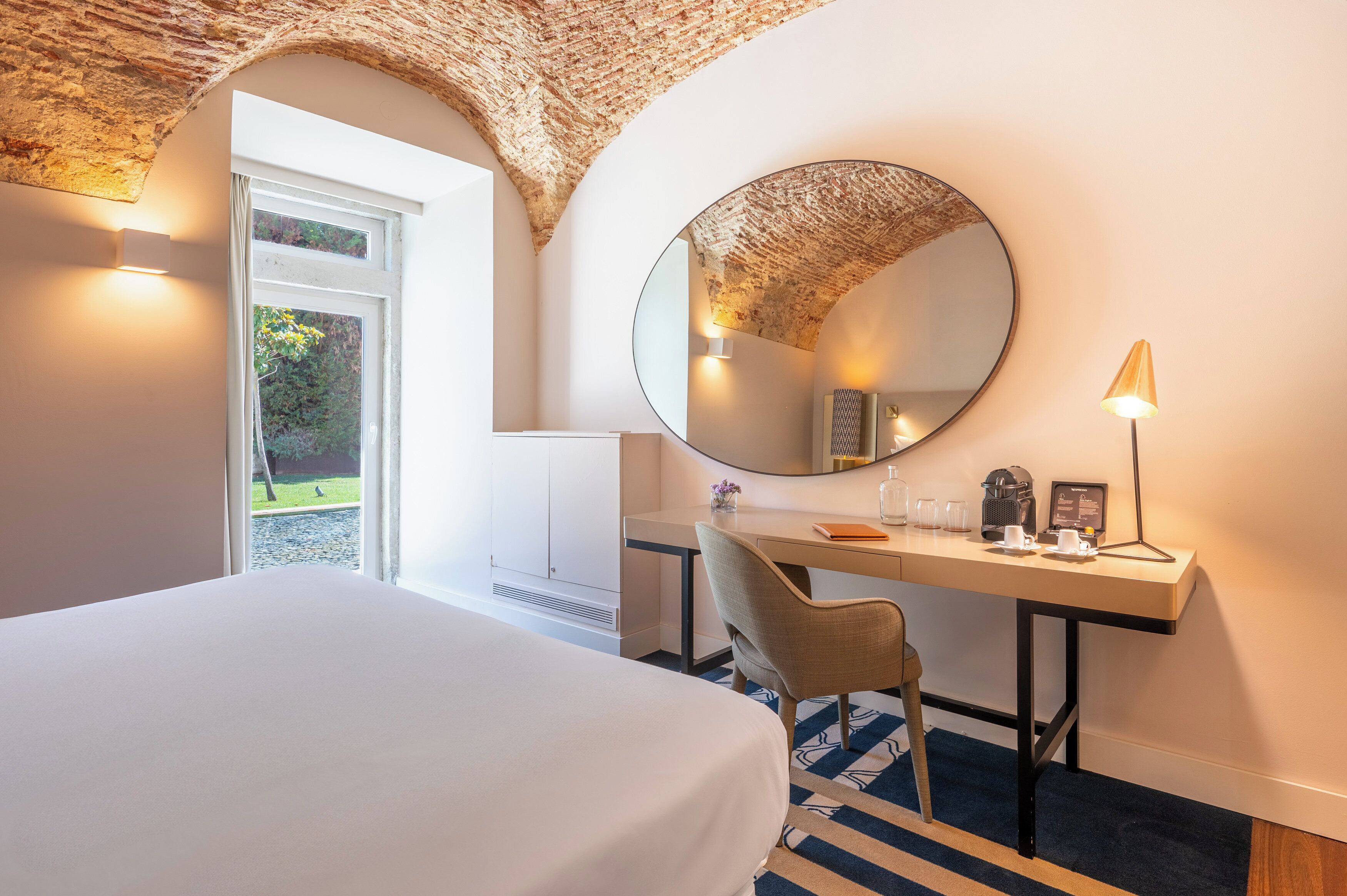 Quarto de hotel com uma cama grande, uma escrivaninha com cadeira, um espelho redondo, uma luminária e vista para o exterior através de uma janela.