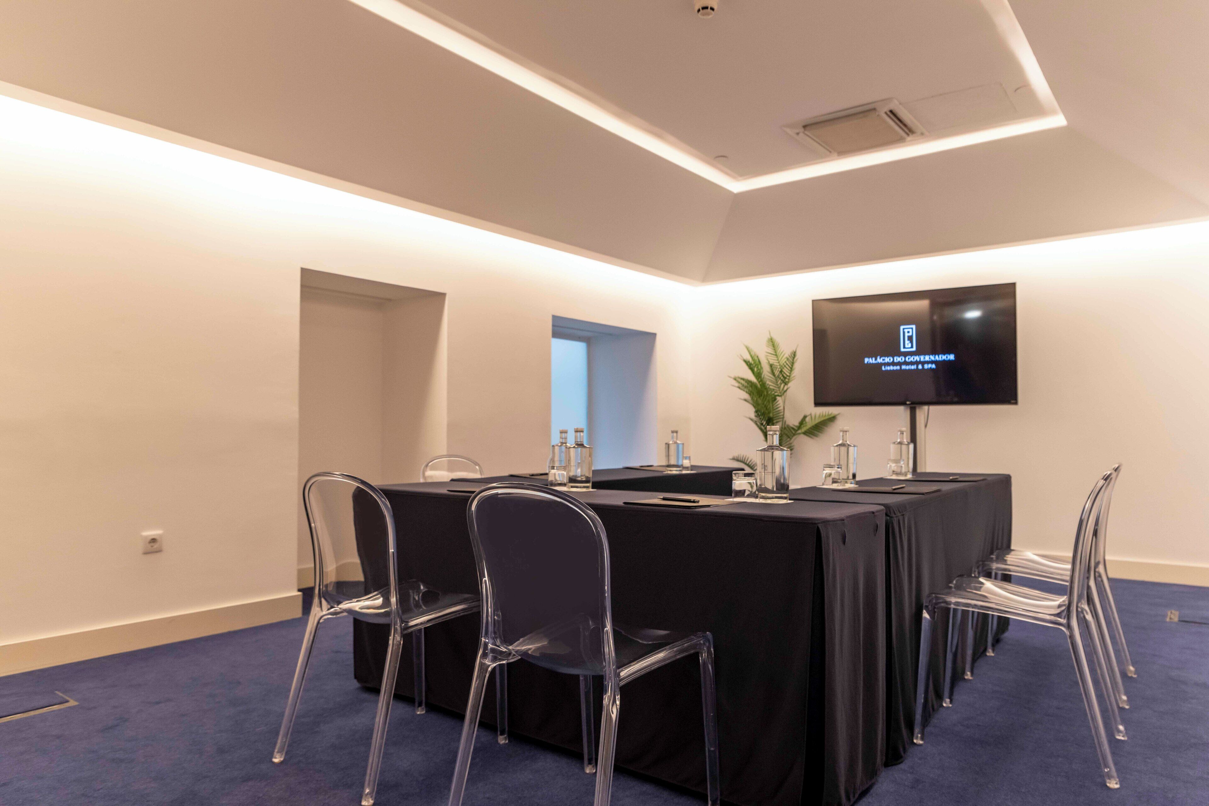 A conference room with a long table, clear chairs, a flat-screen TV, and a potted plant.