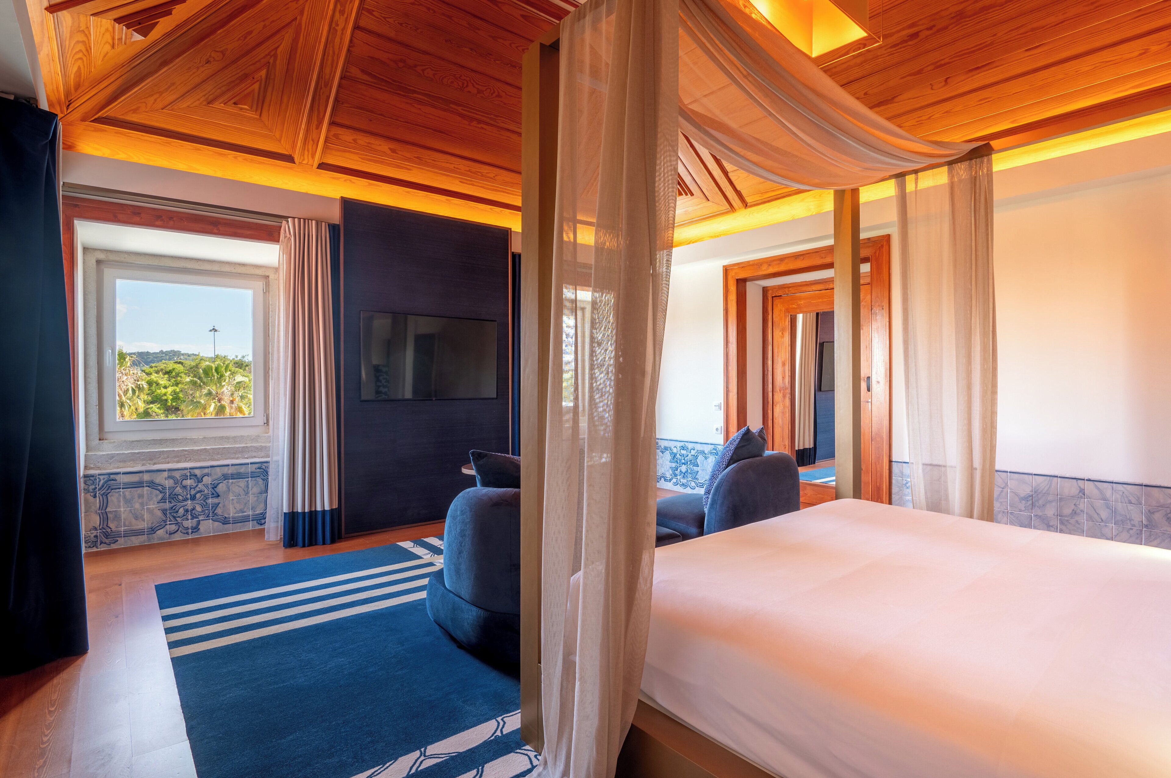 Ein Hotelzimmer mit einem großen Bett, einem Fernseher, Blick auf Grünfläche und einer Holzbalkendecke.