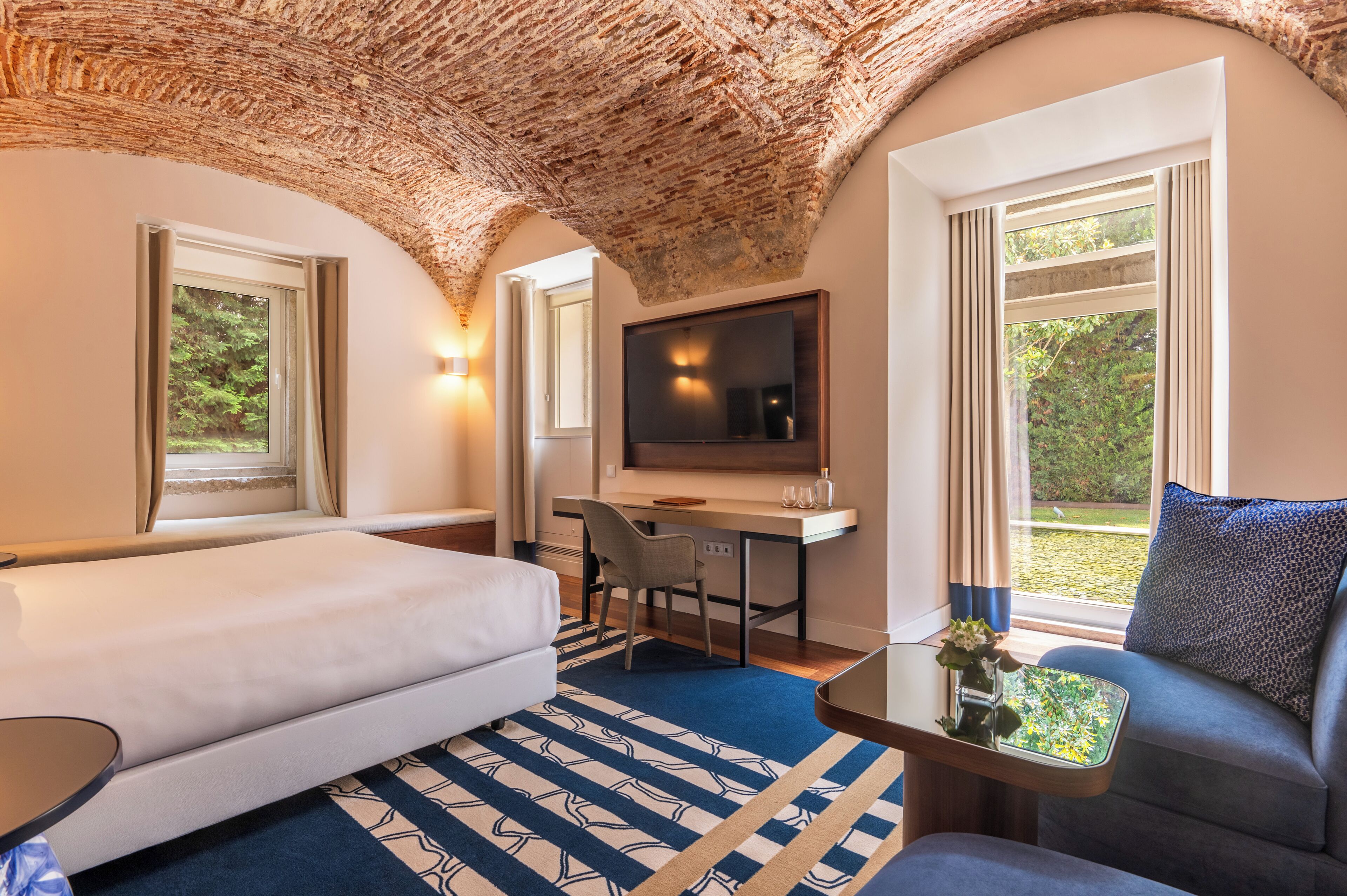 Quarto de hotel com cama, escrivaninha, cadeira, televisão e sofá.