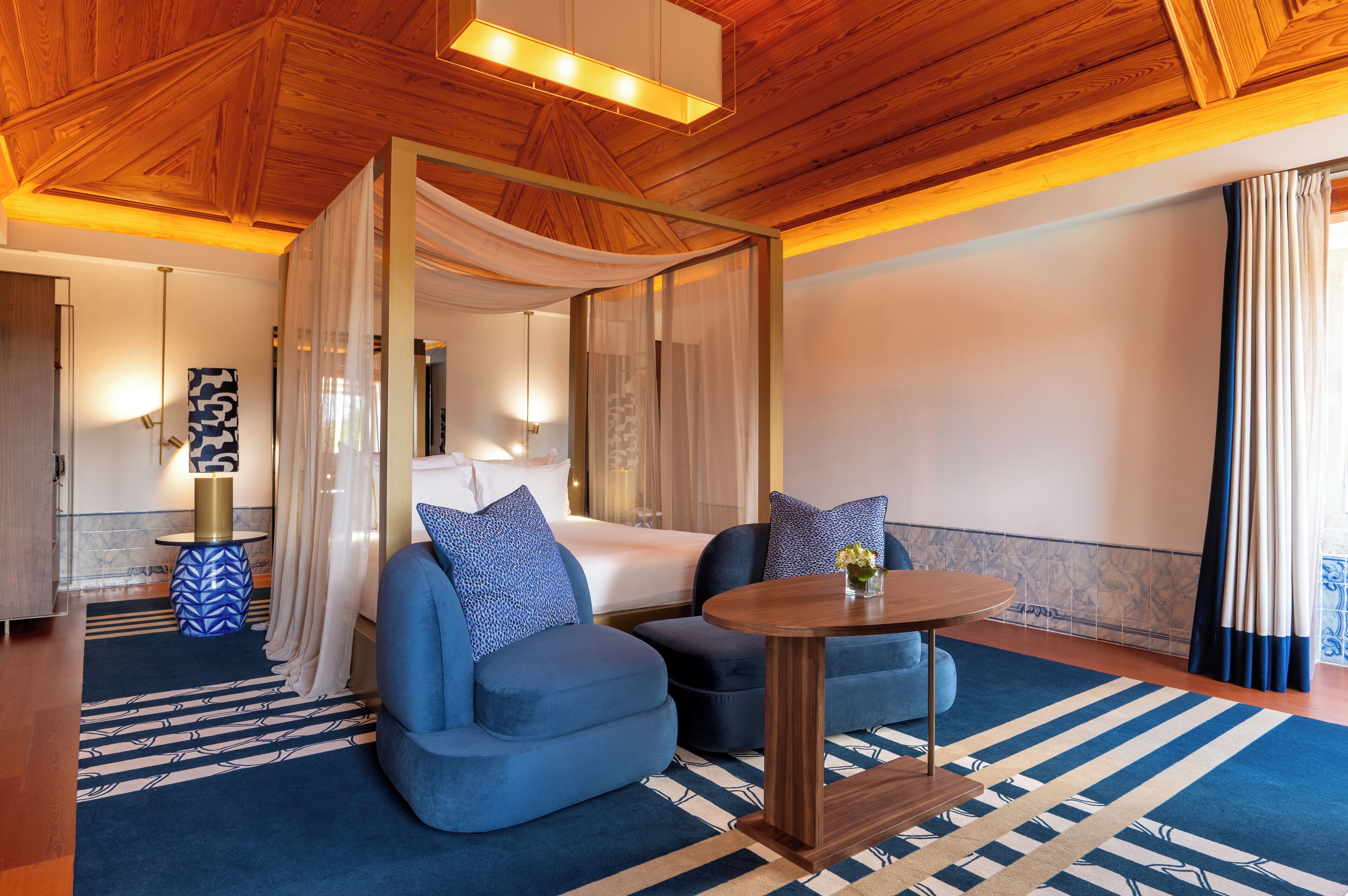 A modern hotel room with a wooden ceiling, a large bed with a canopy, two blue armchairs, a round wooden table, and a patterned rug.