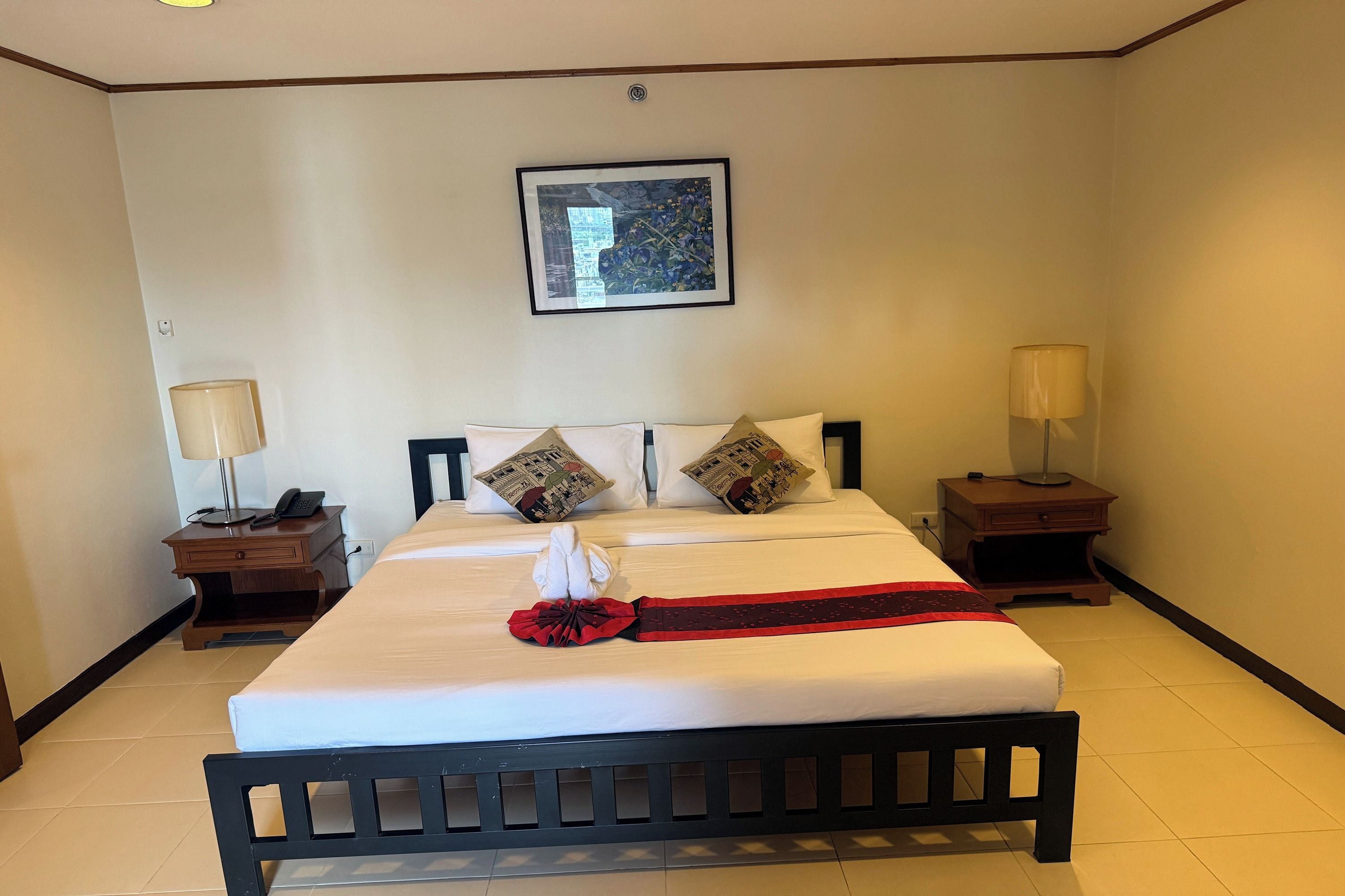 A hotel room with a large bed, two bedside tables, a wall-mounted artwork, and a red decorative item on the bed.