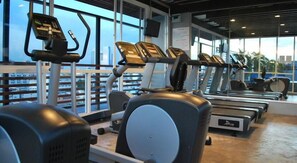 Un gimnasio con cintas de correr y elípticas, amplias ventanas con vista a la ciudad y un balcón con barandilla.