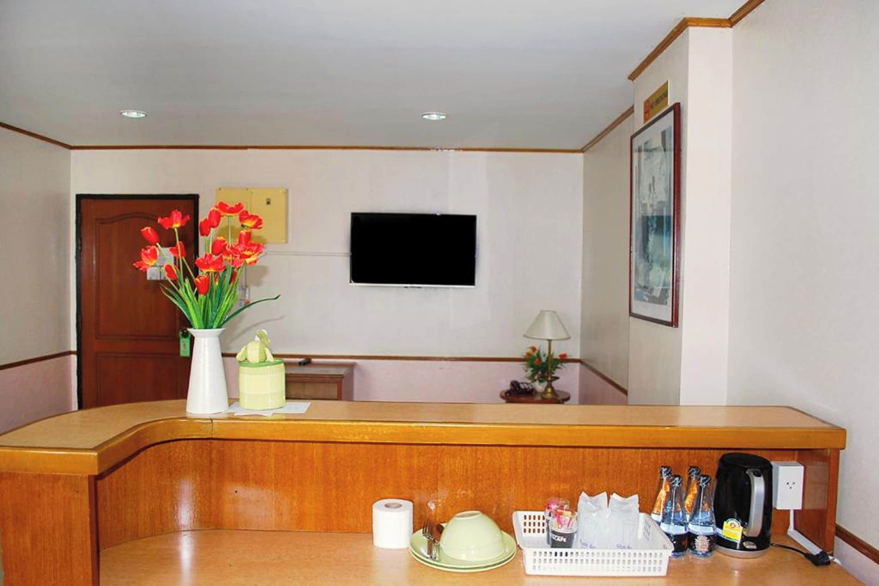 A hotel reception desk with a flat-screen TV mounted on the wall, a wooden reception counter, a vase with red flowers, a small lamp, and a basket with toiletries.
