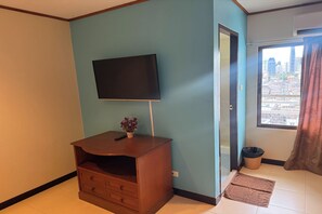 Habitación con televisor de pantalla plana montado en la pared, mueble de entretenimiento de madera con cajones, una planta en maceta y vistas a un paisaje urbano a través de una ventana con cortinas.