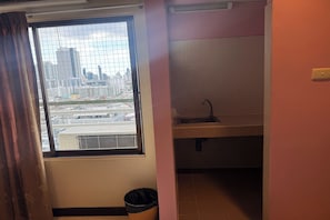 Una pequeña kitchenette con ventanal a una vista urbana, un lavamanos y un cubo de basura.