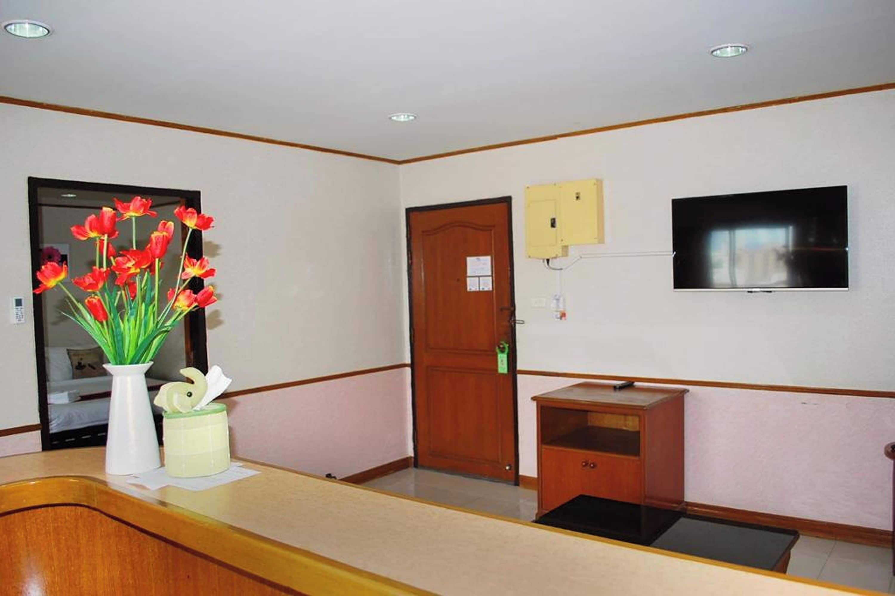 A reception area with a wooden counter, a vase of red flowers, a wooden door, a small wooden cabinet, and a mounted television.