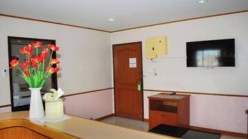 Un área de recepción con mostrador de madera, un jarrón con flores rojas, una puerta de madera, un mueble de madera pequeño y un televisor montado.