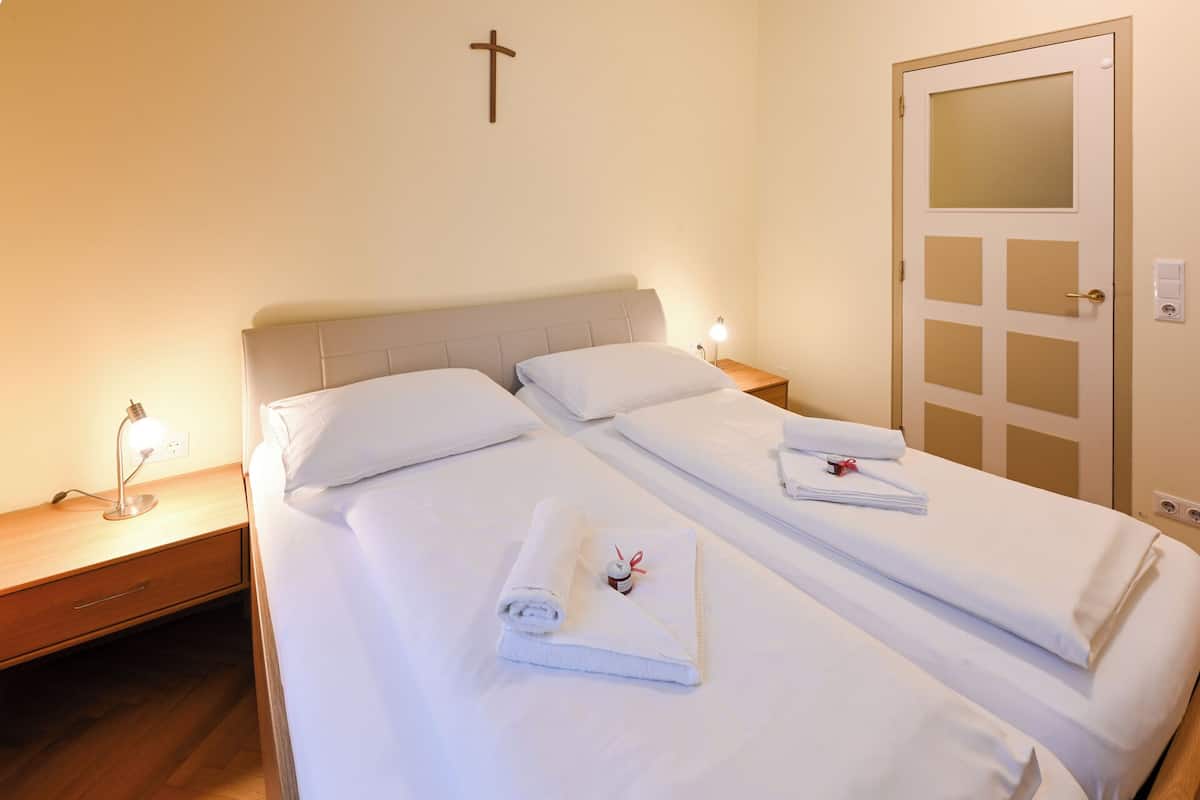 A double bed with white linens, a bedside lamp, a cross on the wall, and a door with a window.